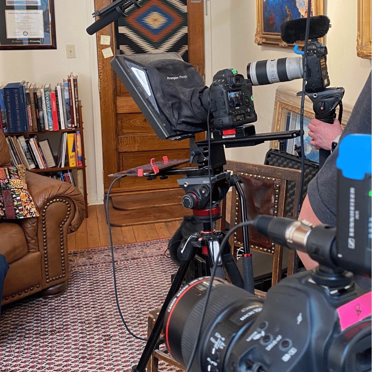 Camera equipment set up on a tripod inside a room with bookshelves, framed artwork, and a leather armchair.
