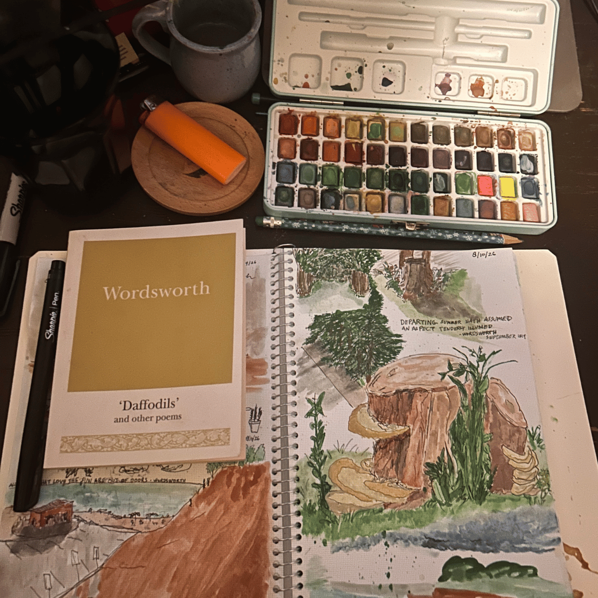 An art workspace with a watercolor painting of a tree stump with greenery, a watercolor palette, a sketchbook, a book titled 'Wordsworth Daffodils and other poems,' a black Sharpie pen, a paintbrush, a ceramic mug, a small circular wooden platform, and an orange lighter.