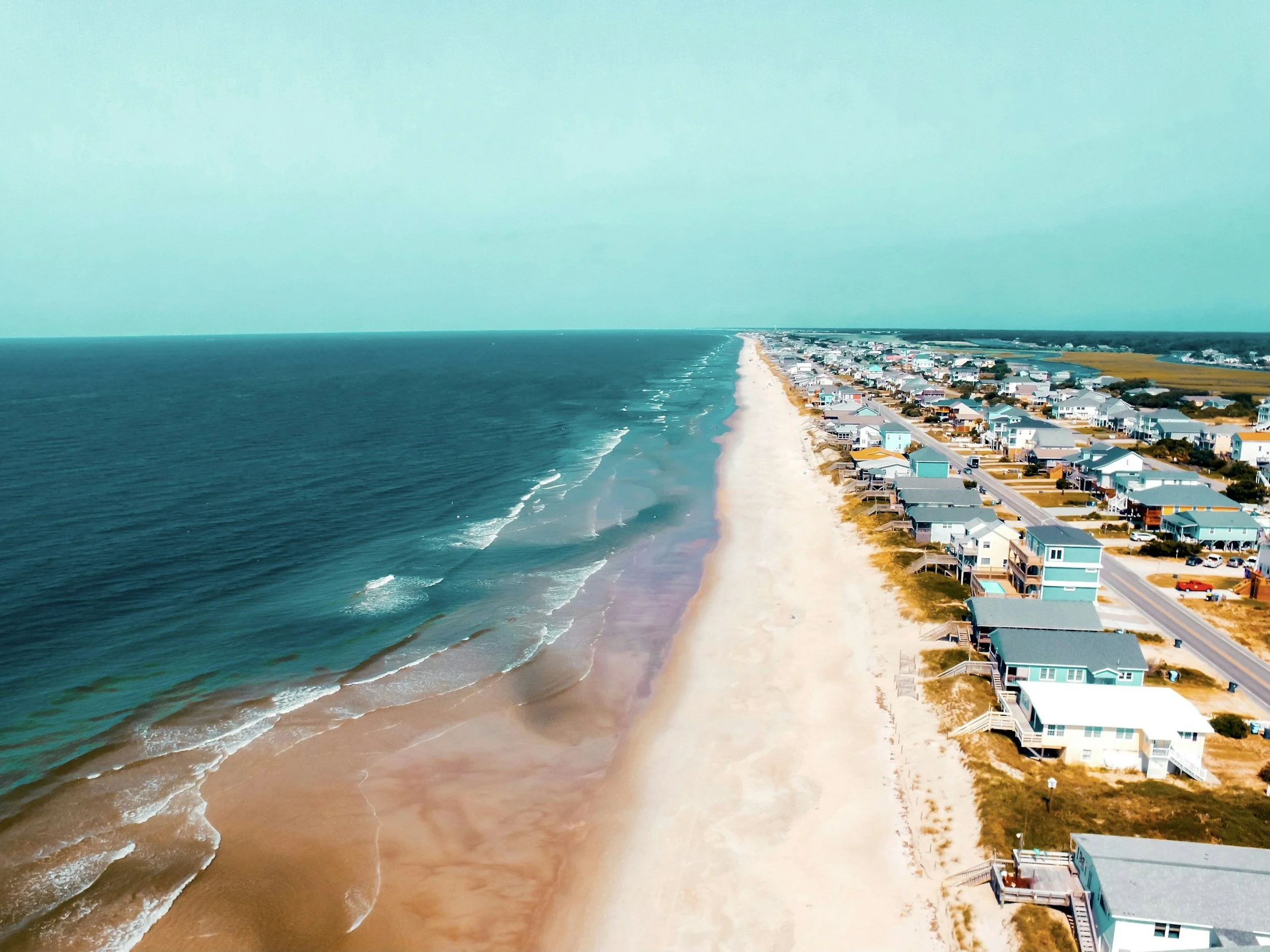 An aerial view of a beach with turquoise water, white sand, and a row of colorful beach houses along the shoreline under a clear sky.