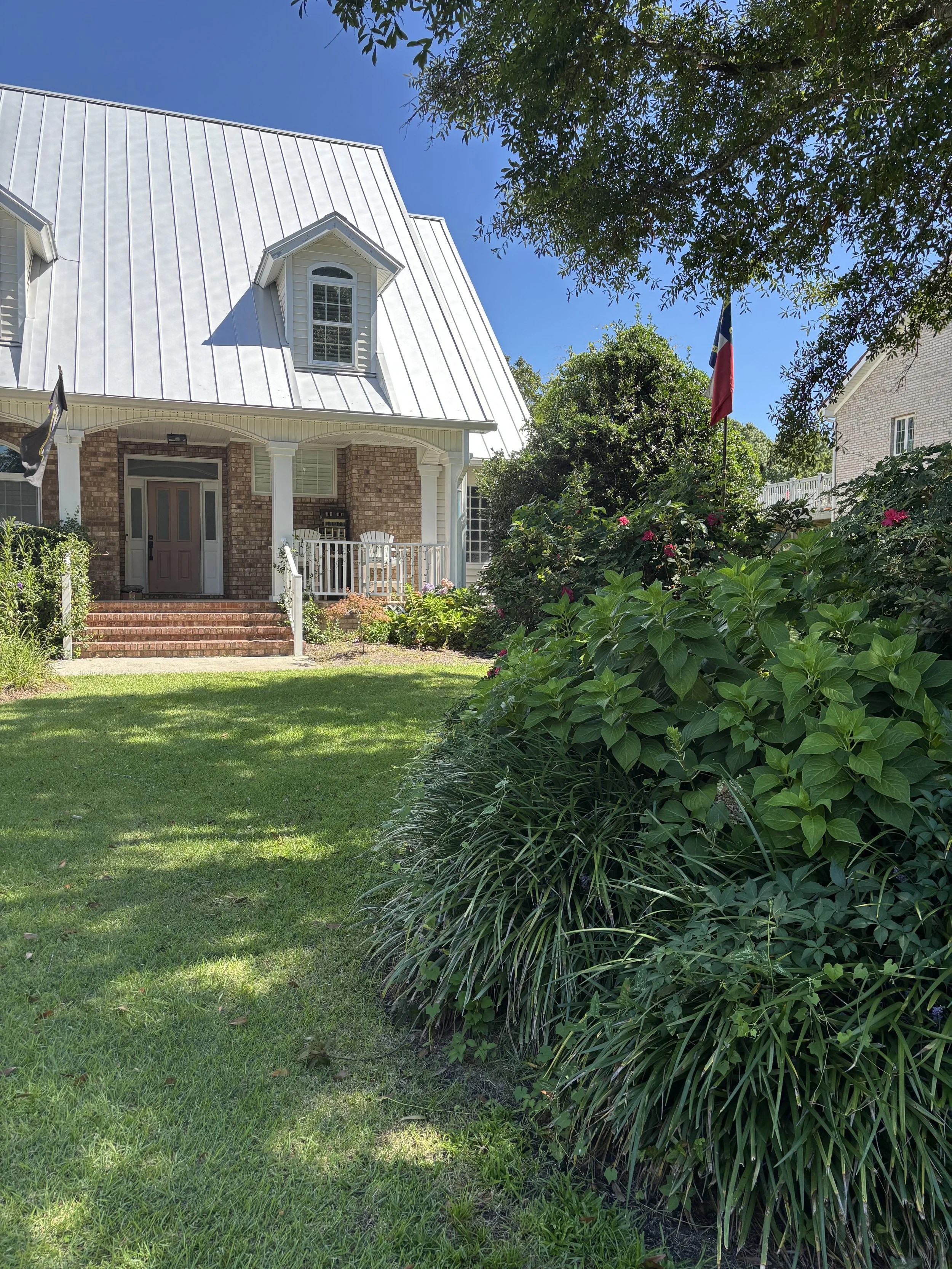 Front yard of a house with a porch, brick steps, lush green lawn, and large bushes, with a tree and American flag in the yard under a clear blue sky.