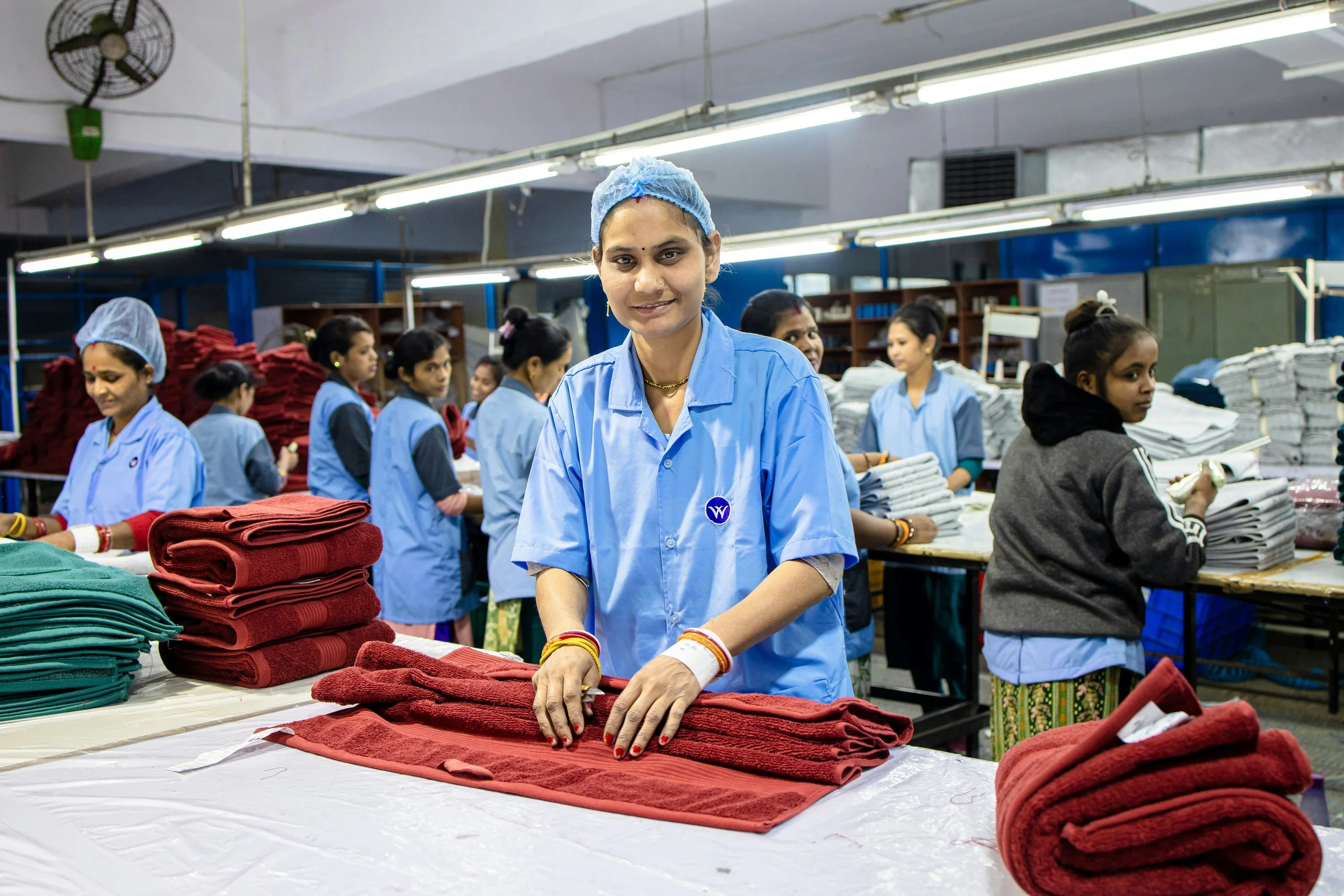 A worker folds a red towel at a factory table while other workers handle stacks of towels behind her