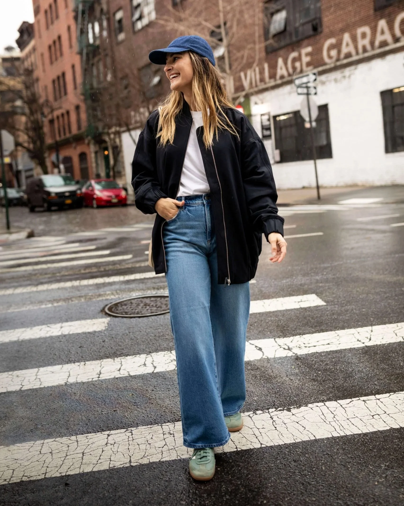 A woman crossing a New York City street in a casual fall outfit of jeans, a jacket, and a baseball cap