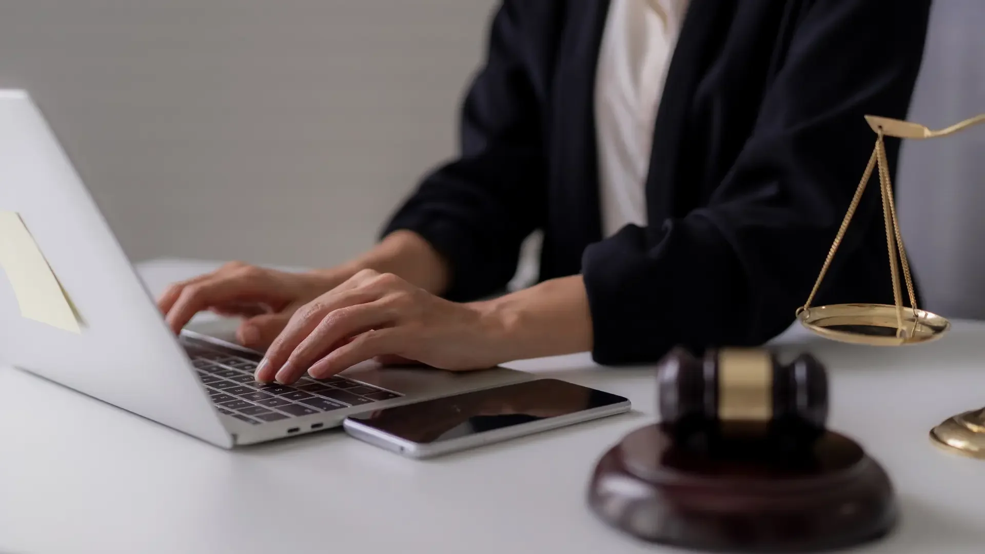 Person wearing a dark jacket typing on a laptop at a desk with legal scales, a gavel, and a smartphone.