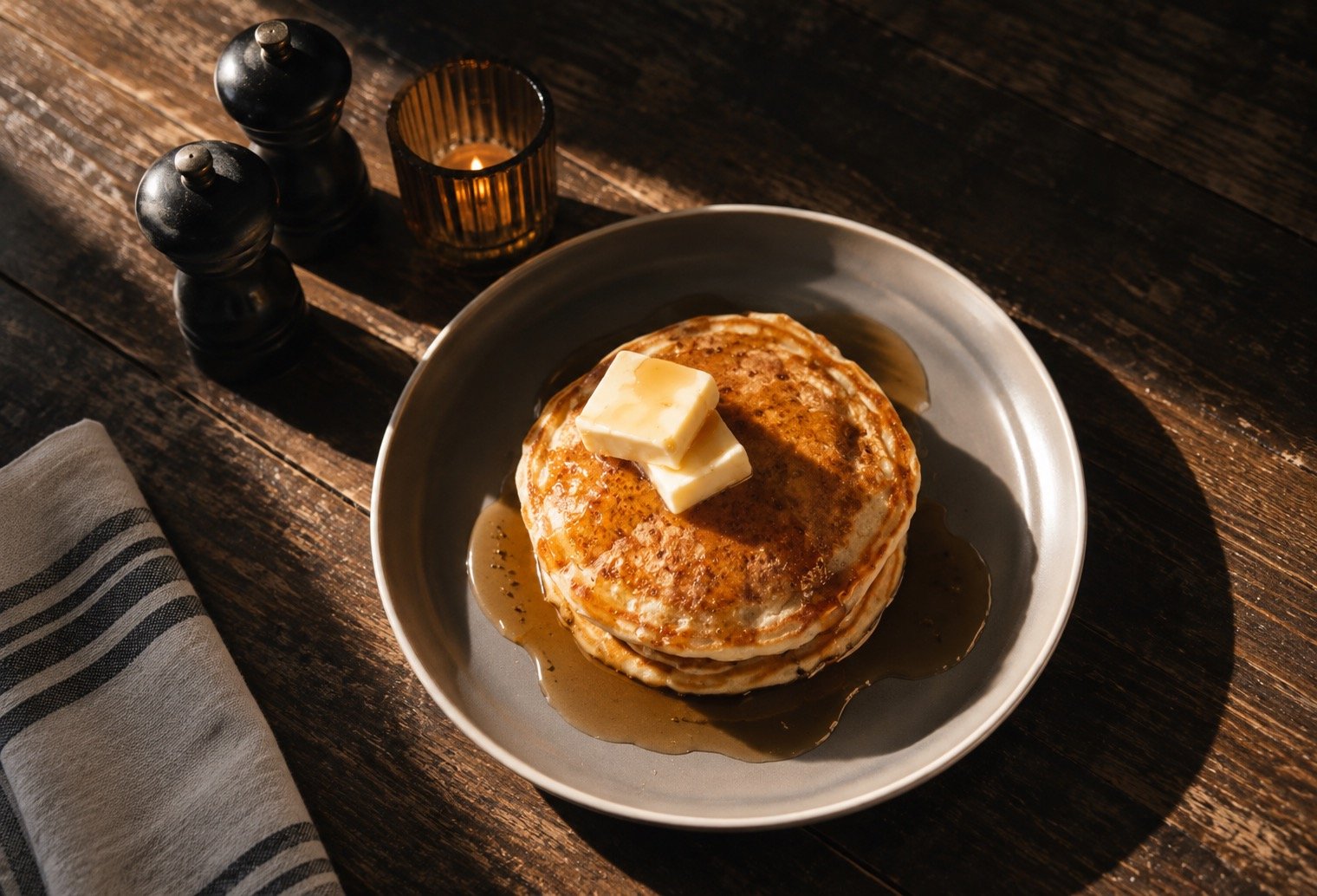 Stack of pancakes topped with butter and syrup on a gray plate, with salt and pepper shakers, a candle, and a napkin nearby on a wooden table.