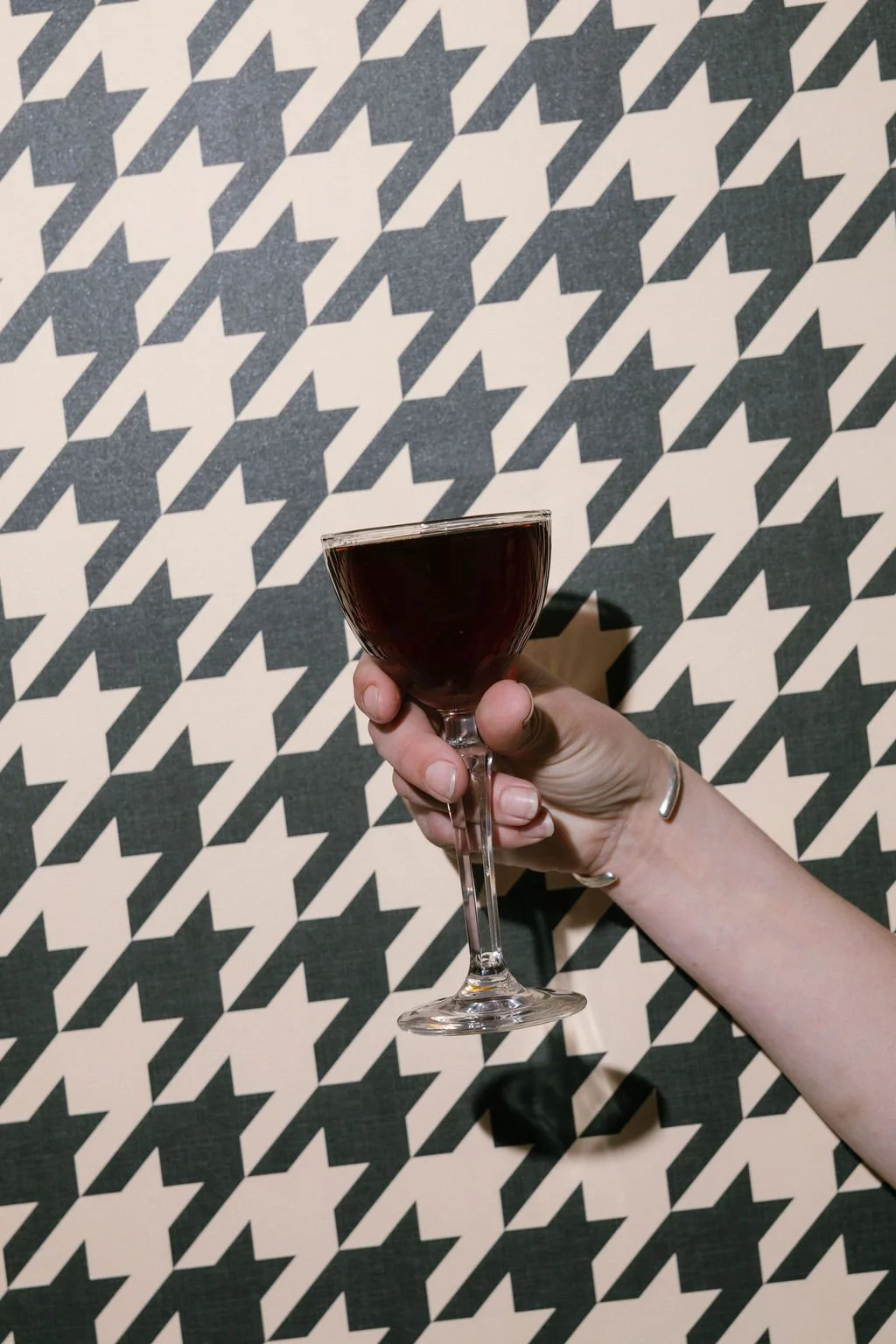 A person holding a glass of dark red wine against a black and white houndstooth patterned background.