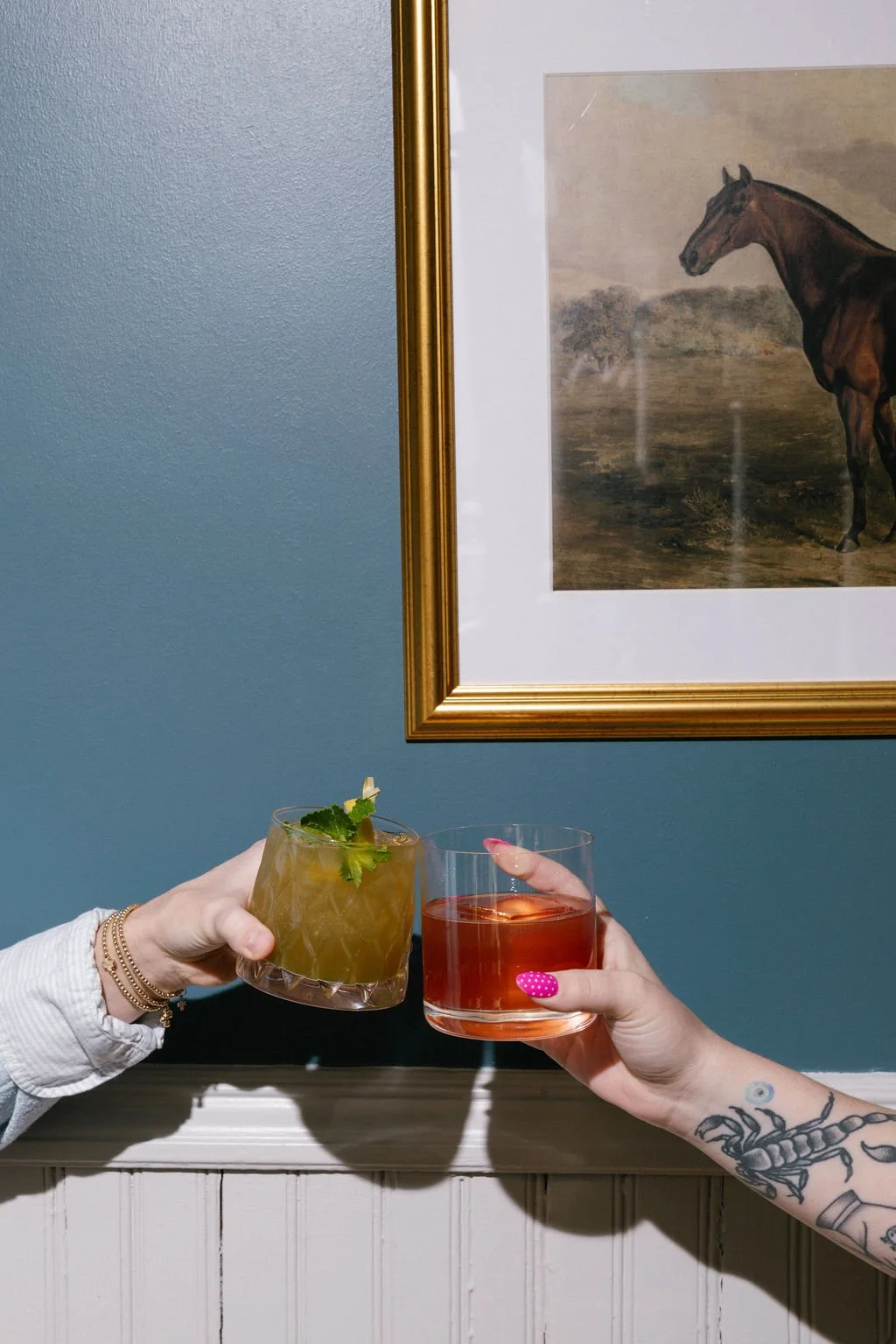 Two people toasting with cocktails, one with a mint garnish and the other in a clear glass, against a blue wall with a horse painting and gold frame.