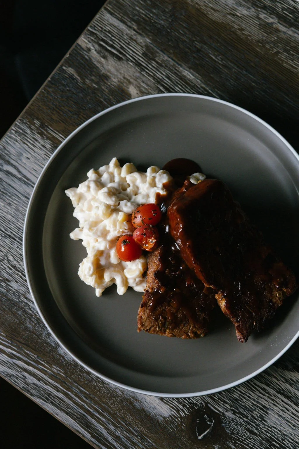Cooked ribs with barbecue sauce, served with macaroni and cheese, and cherry tomatoes on a gray plate.