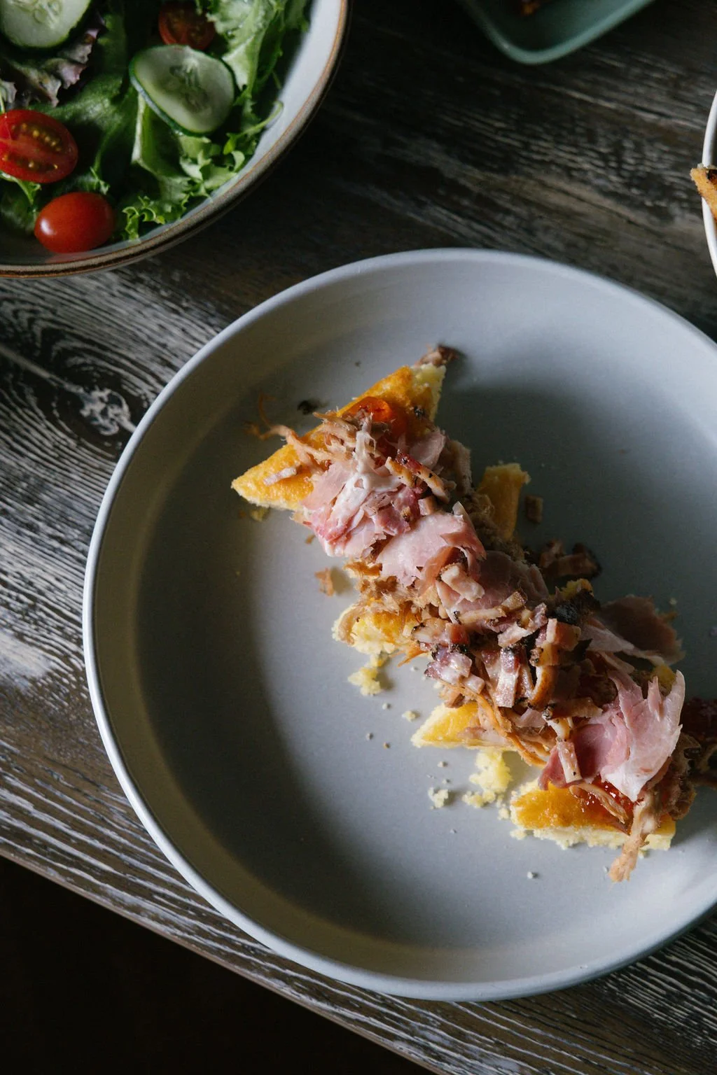 A partially eaten slice of pizza with ham and melted cheese on a white plate, and a bowl of mixed salad with cherry tomatoes and cucumber slices on a wooden table.