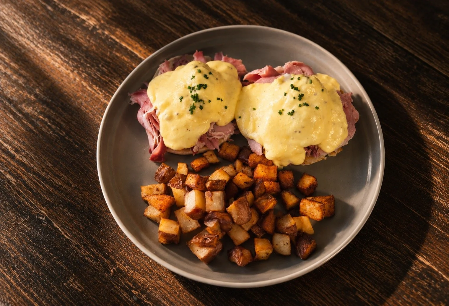 Plate of Eggs Benedict with ham and hollandaise sauce, served with roasted breakfast potatoes on a dark wooden table.