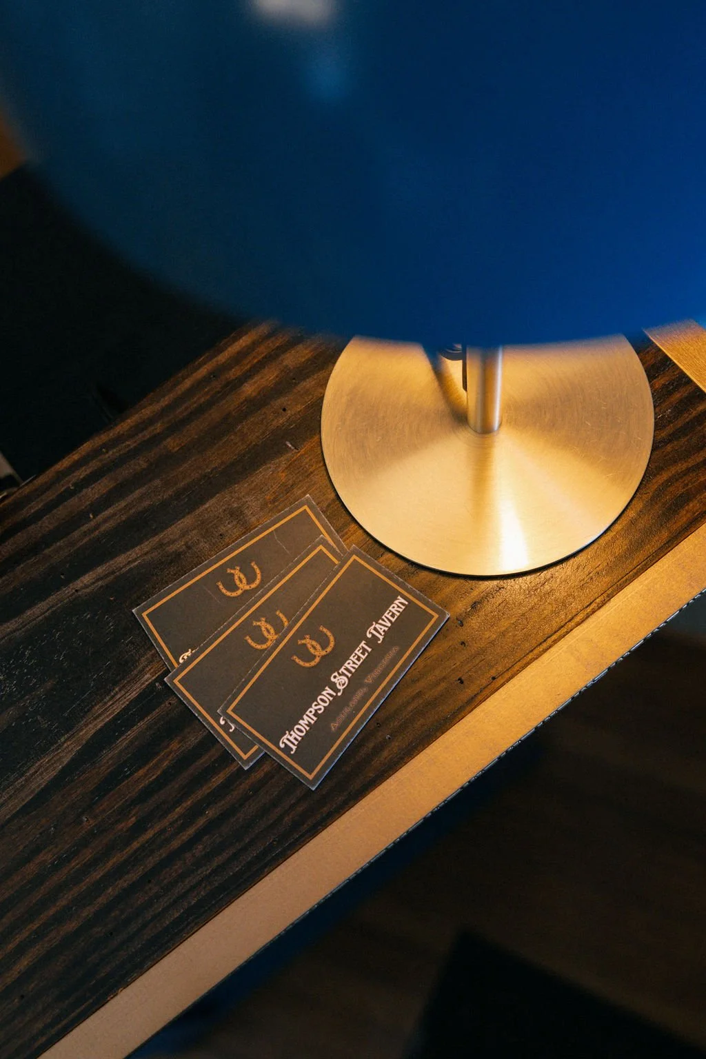 Black cards with a gold horseshoe emblem and the text 'Thompson Street Tavern' on a dark wooden table next to a lamp with a circular metal base and blue shade.