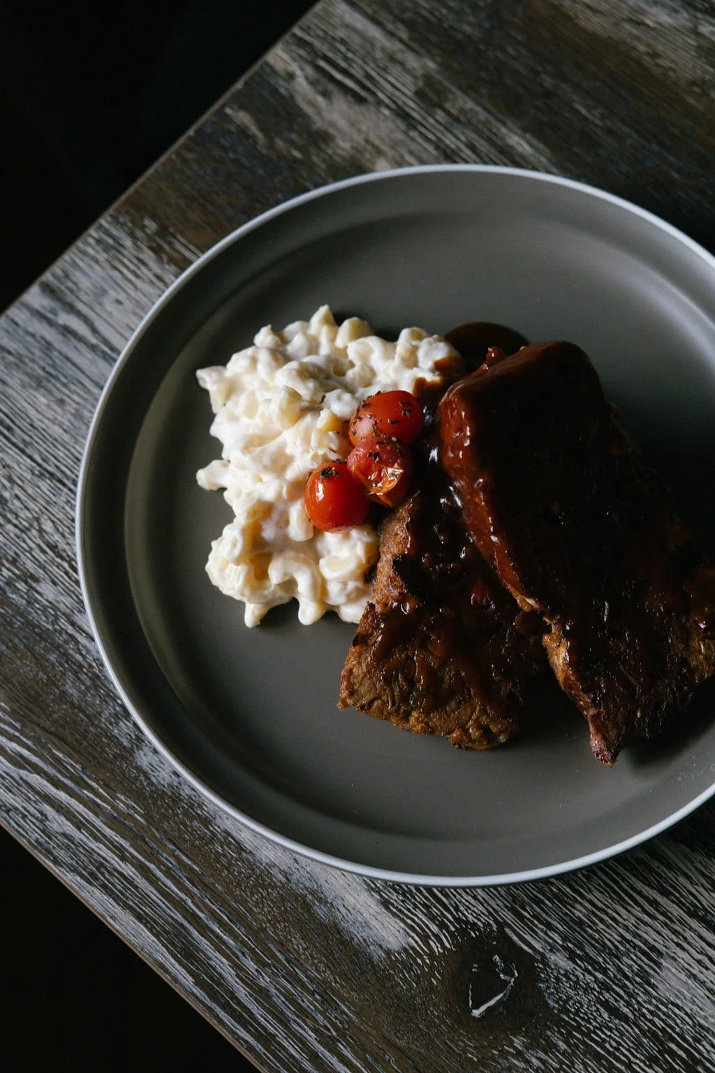 Plate of macaroni and cheese with cherry tomatoes, two braised beef short ribs with sauce, all on a dark gray plate on a wooden table.
