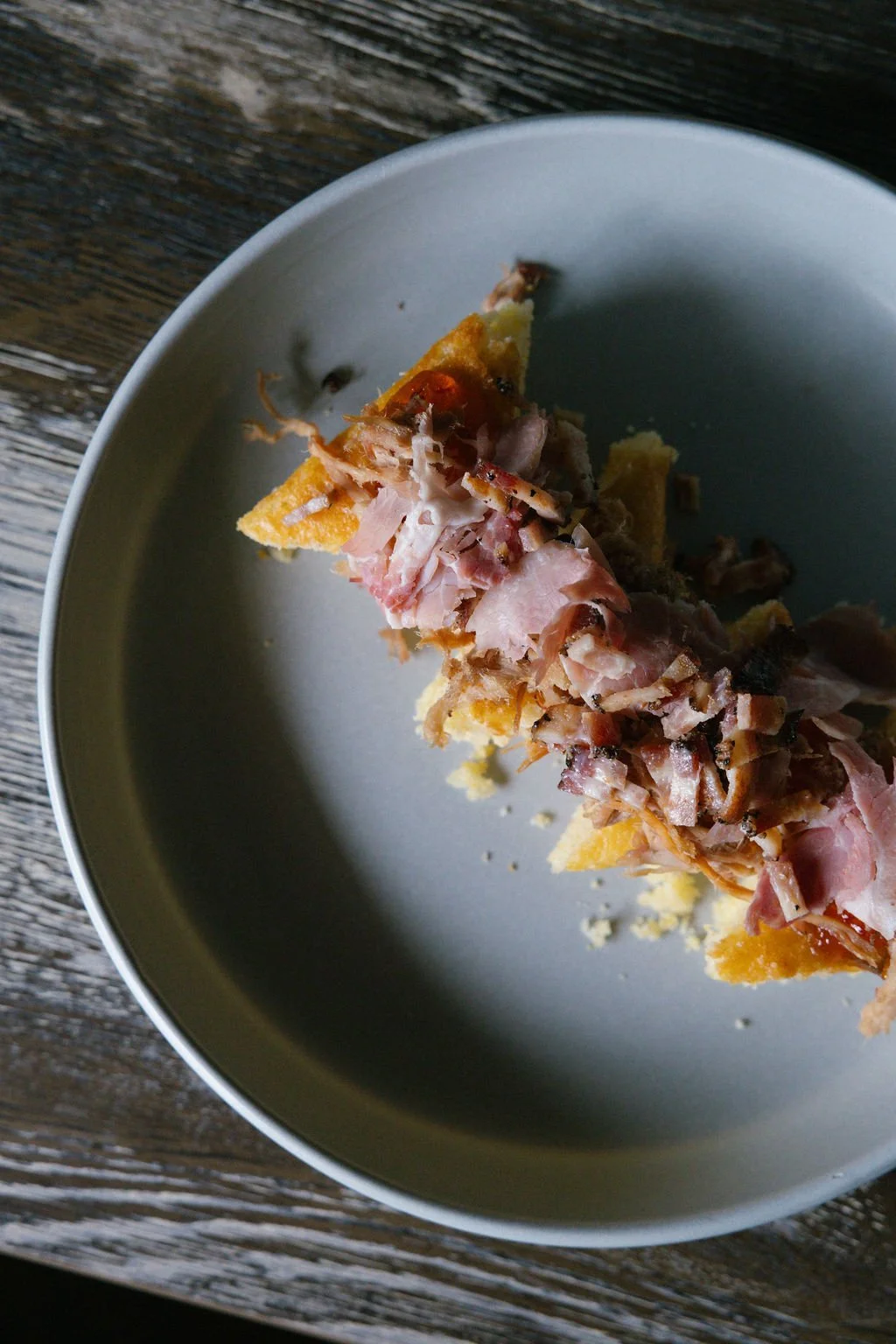 A slice of pie with ham, bacon, and cheese on a white plate resting on a wooden surface.