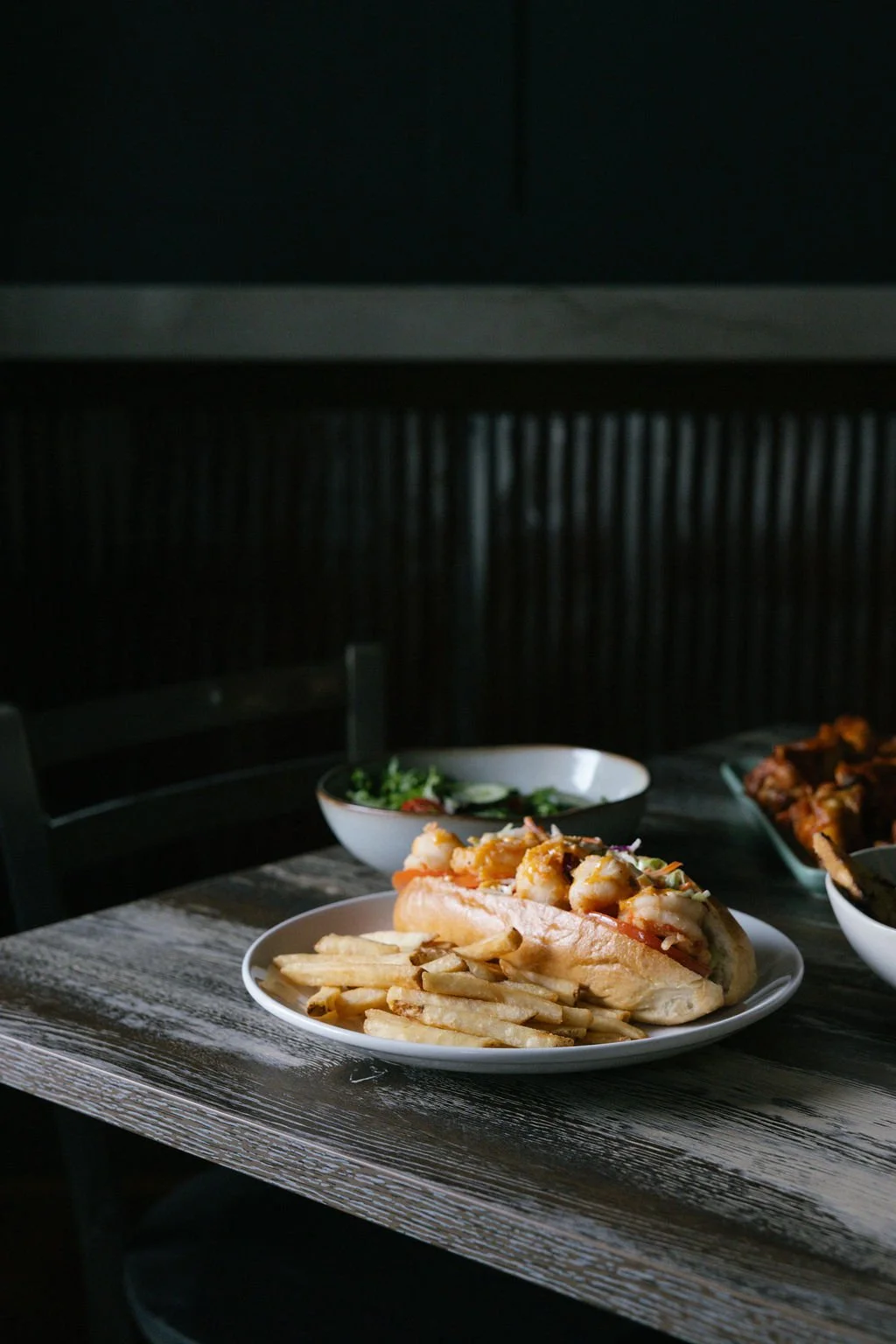 A plate with a sandwich filled with shrimp on a wooden table, with French fries on the side, and two bowls of salad in the background.