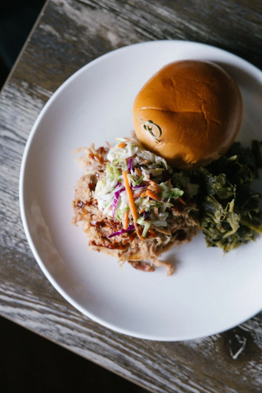 Barbecue pulled pork sandwich with coleslaw on a white plate, accompanied by a bun and a small serving of greens, on a rustic wooden table.