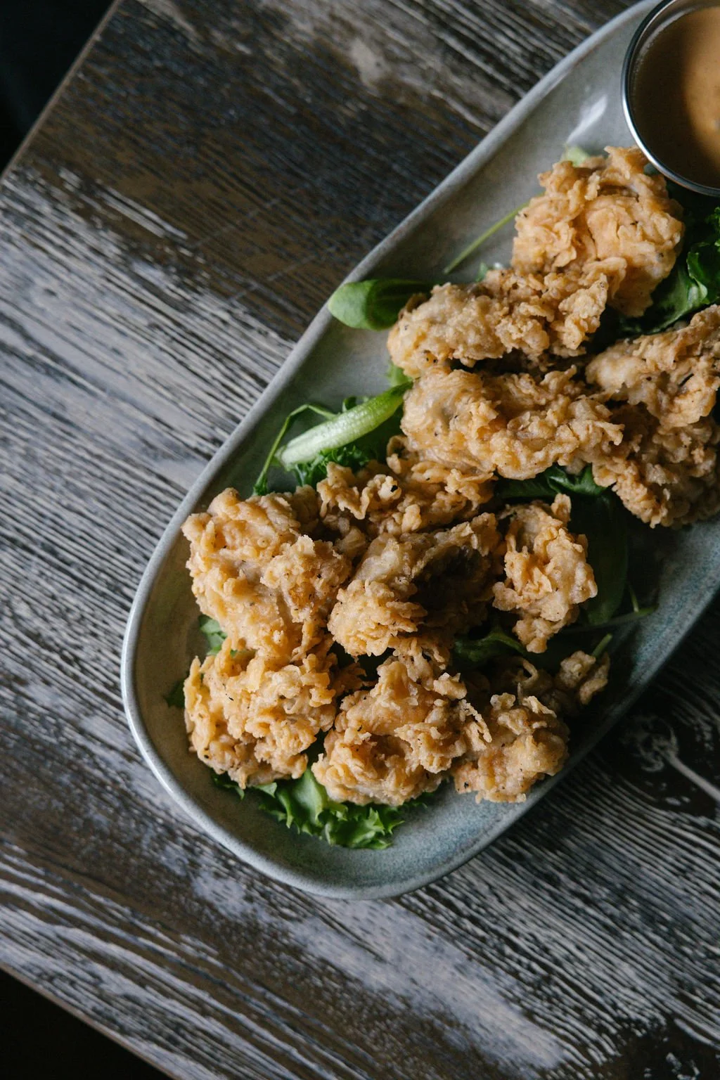 Fried chicken tenders on a bed of fresh greens on a ceramic platter with a small bowl of dipping sauce, placed on a wooden table.