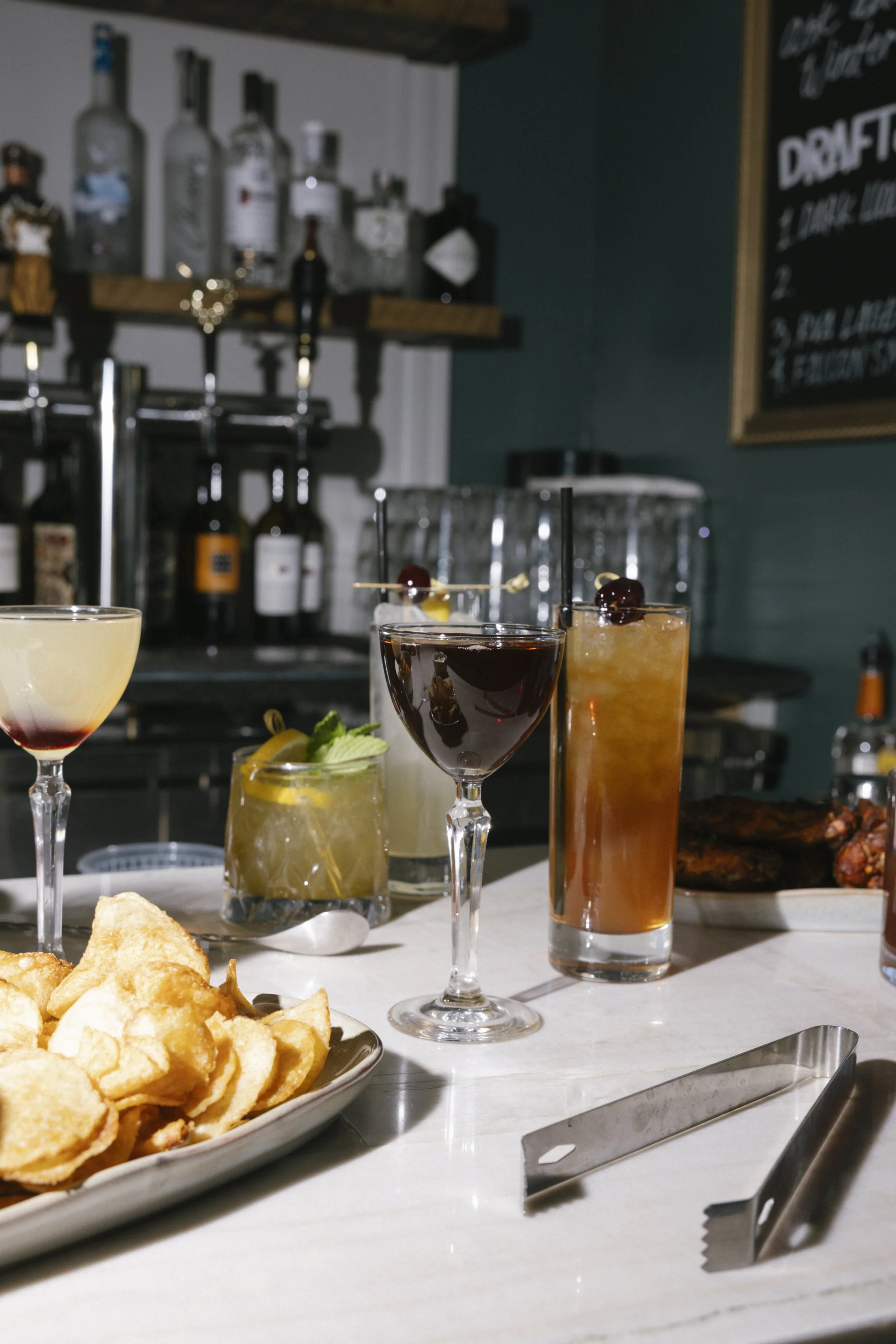 A bar or restaurant setting with various colorful cocktails garnished with fruit and cherries, a plate of fried snack chips, liquor bottles on a shelf in the background, and a menu on the wall.