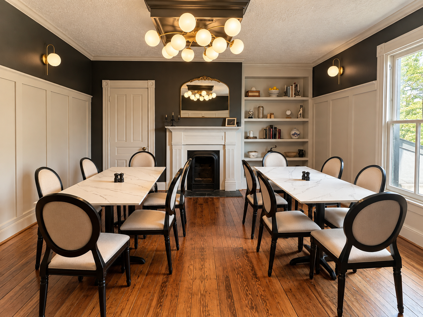 A dining room with two white marble-top tables and eight beige upholstered chairs with black frames, a white fireplace, built-in shelves with decor, dark gray walls, wood flooring, wall sconces, a ceiling light fixture, and a large window.
