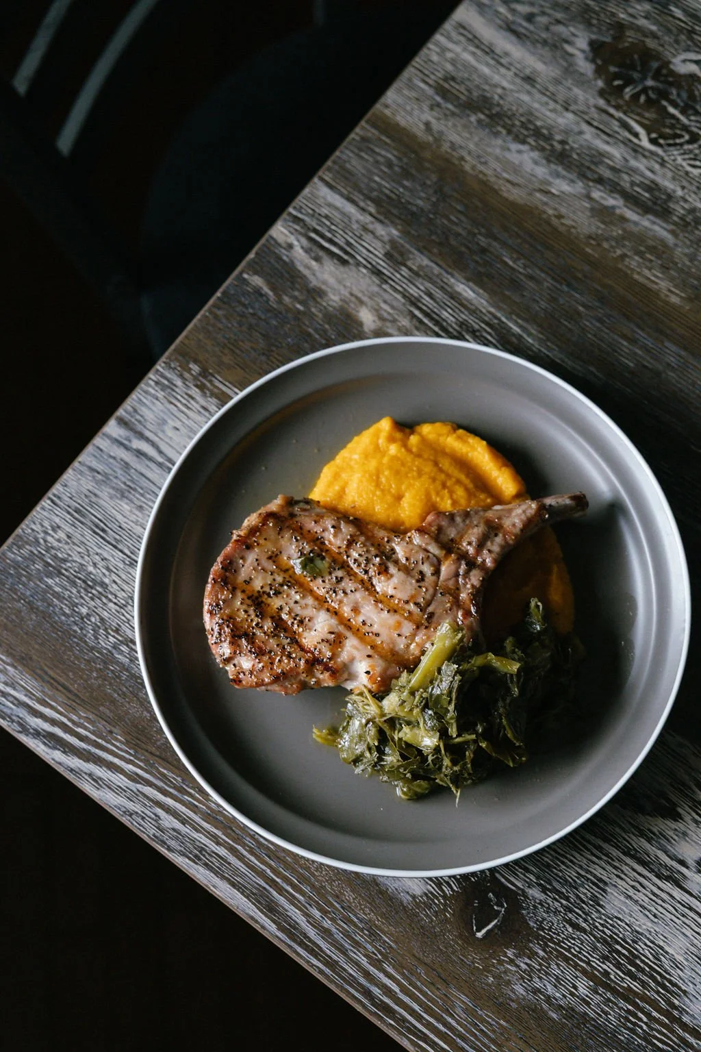 Grilled pork chop with mashed sweet potatoes and cooked greens on a gray plate, on a rustic wooden table.