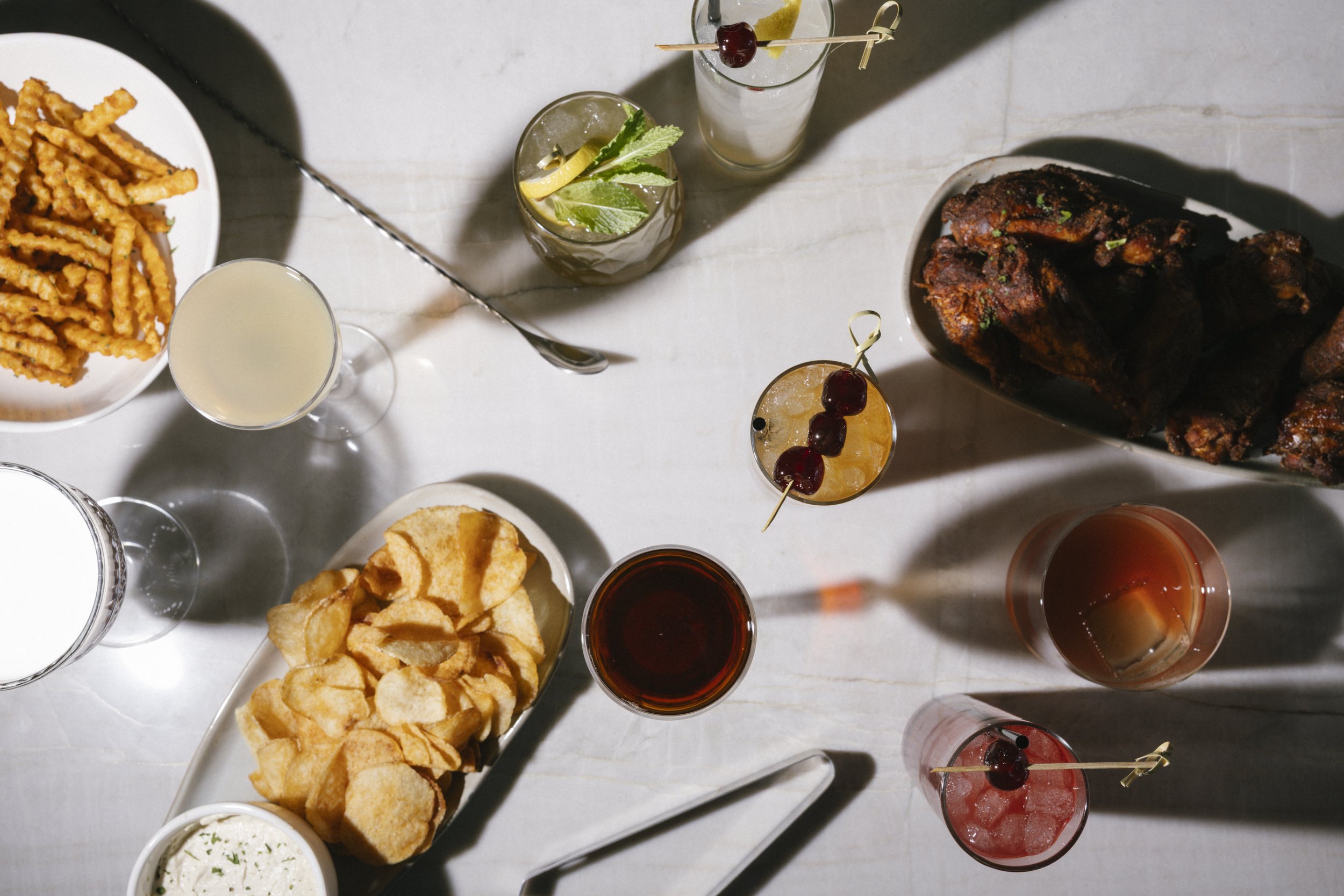 Top-down view of a table with bar drinks, a plate of curly fries, a bowl of potato chips, some fried snacks, and a bowl of creamy dip.