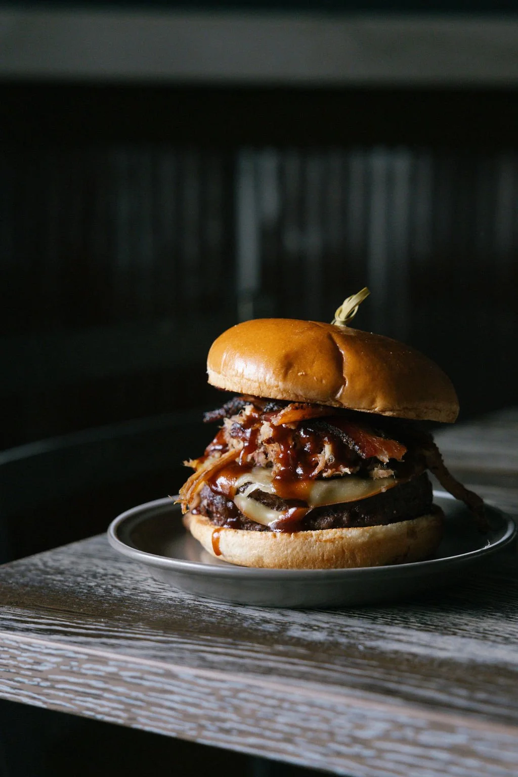 Barbecue burger with beef patty, melted cheese, pulled pork, and barbecue sauce in a bun, served on a small plate on a rustic wooden table.
