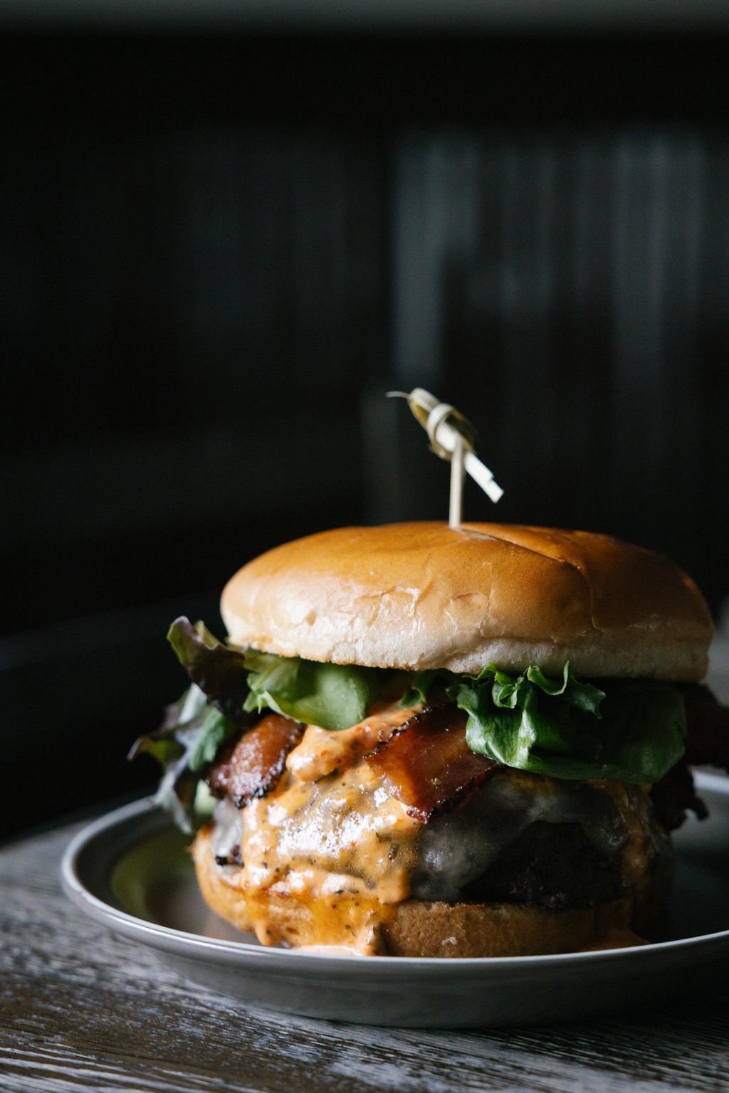 Close-up of a gourmet burger with lettuce, bacon, melted cheese, and sauce, on a plate.