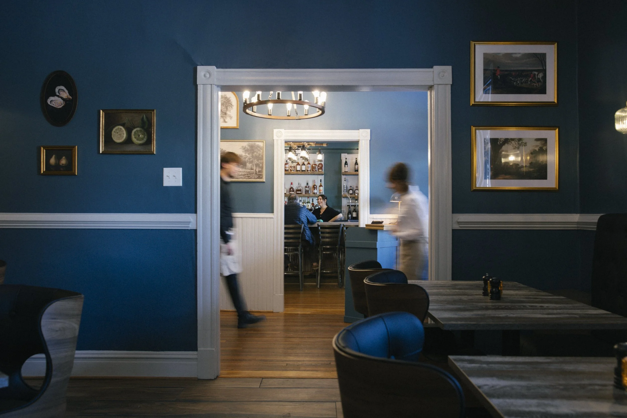 Interior view of a restaurant with blue walls, decorated with framed artwork and photos. The foreground shows black chairs and dark wooden tables, some of which have small condiment bottles. In the background, a doorway reveals a bar area with shelves of liquor bottles and a person sitting and talking with another person, with a chandelier hanging overhead. Two servers are moving through the scene, appearing blurred.