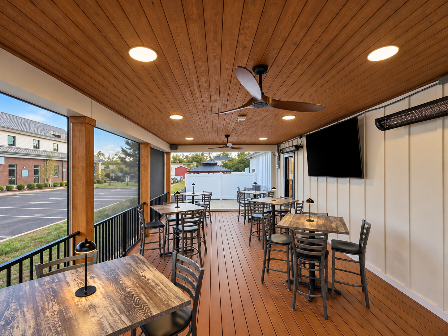 Indoor patio with wooden flooring and ceiling, several tables with chairs, ceiling fans, large wall-mounted TV, large windows showing parking lot and buildings outside.