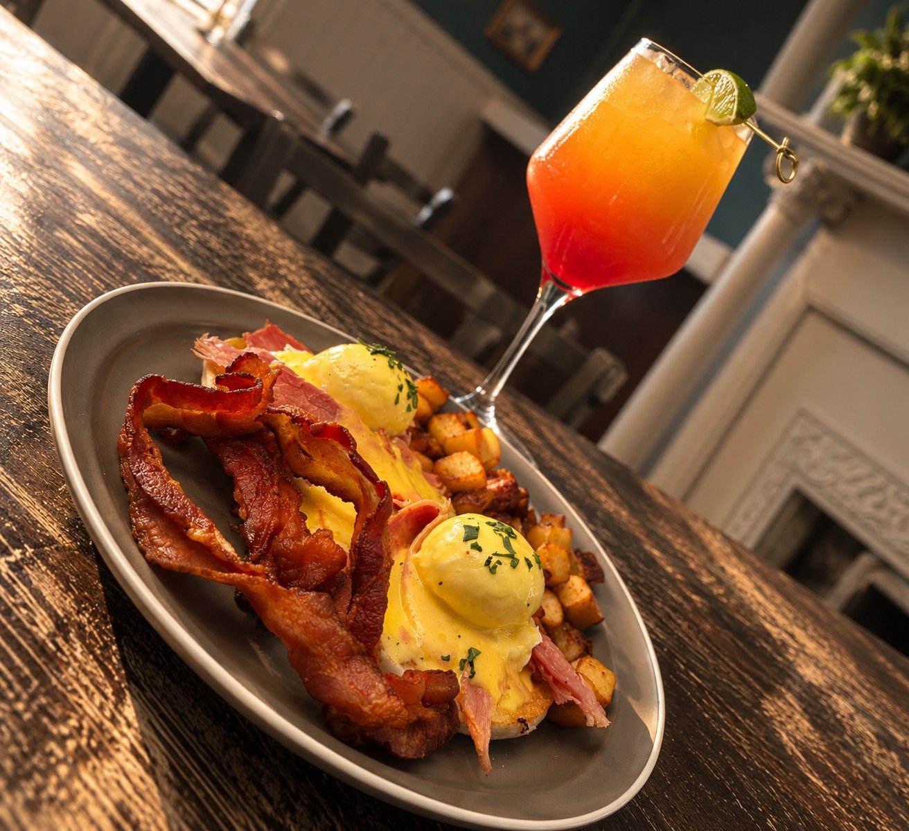 A plate of breakfast food including bacon, poached eggs, and roasted potatoes, with a colorful cocktail garnished with lime on a wooden table.