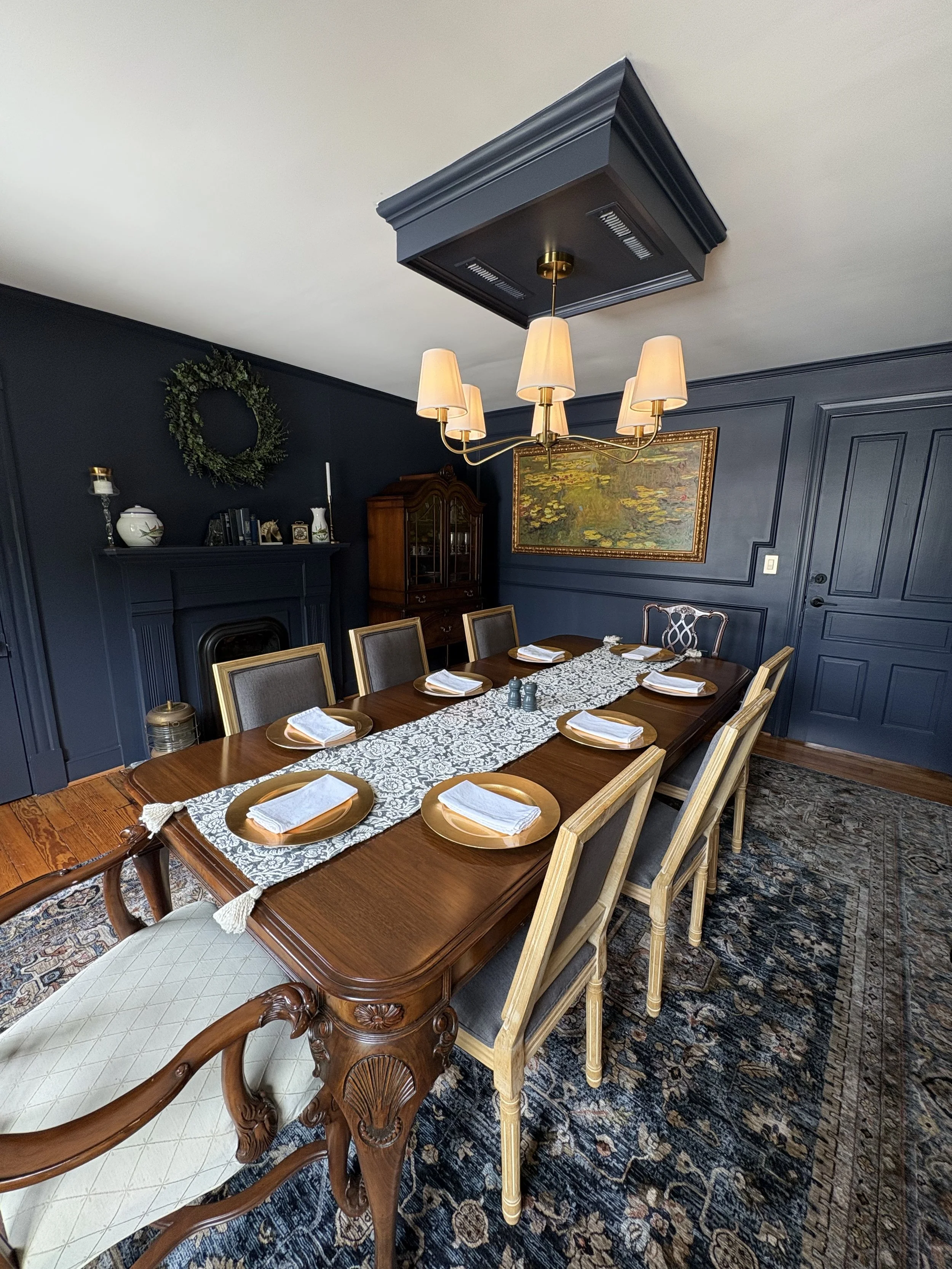 A dining room with a large wooden table set with plates, napkins, and a white lace table runner. The room has dark blue paneled walls, a chandelier with six beige lampshades, a framed painting of water lilies, and a dark wood china cabinet. The carpet is a patterned dark rug, and there is a white cushioned armchair at the end of the table.