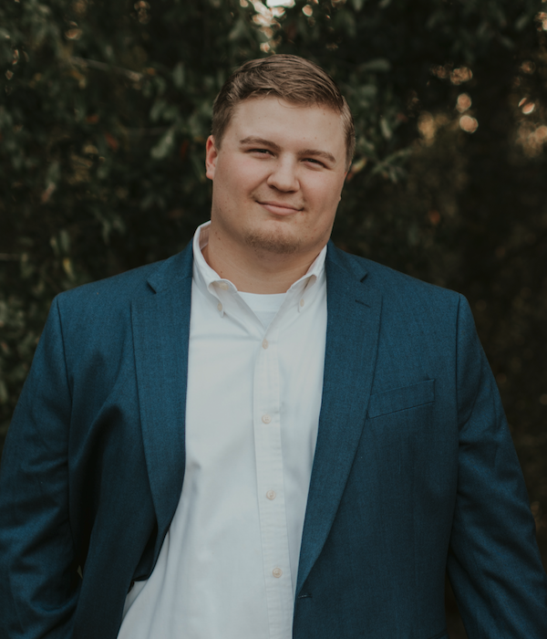 A young man with short brown hair wearing a white button-up shirt and a blue blazer, standing outdoors in front of green foliage.