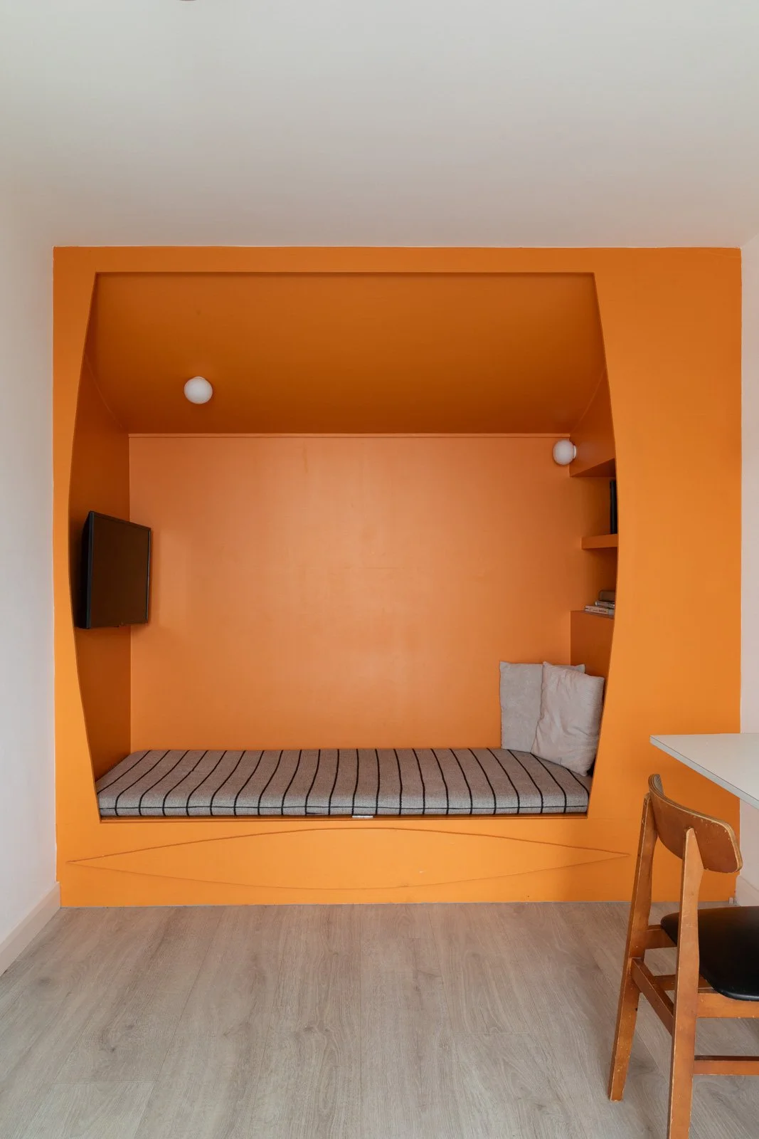 Room with orange walls, built-in sofa bed with striped mattress, TV on the left, pillows on the right, and a small shelf with books on the right side, wooden chair in foreground, and pendant lights on ceiling.