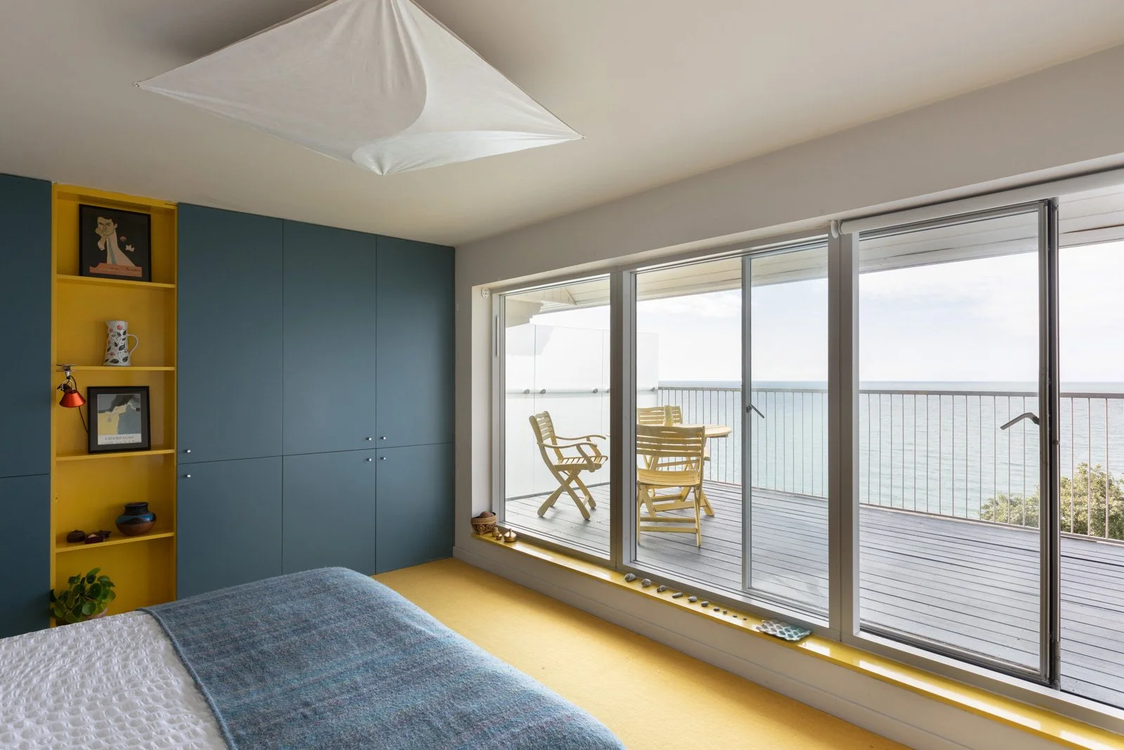 A bedroom with large sliding glass doors leading to a balcony overlooking the sea. The room has blue cabinets, a yellow shelf with decorations, a bed with white and blue bedding, and a ceiling light.