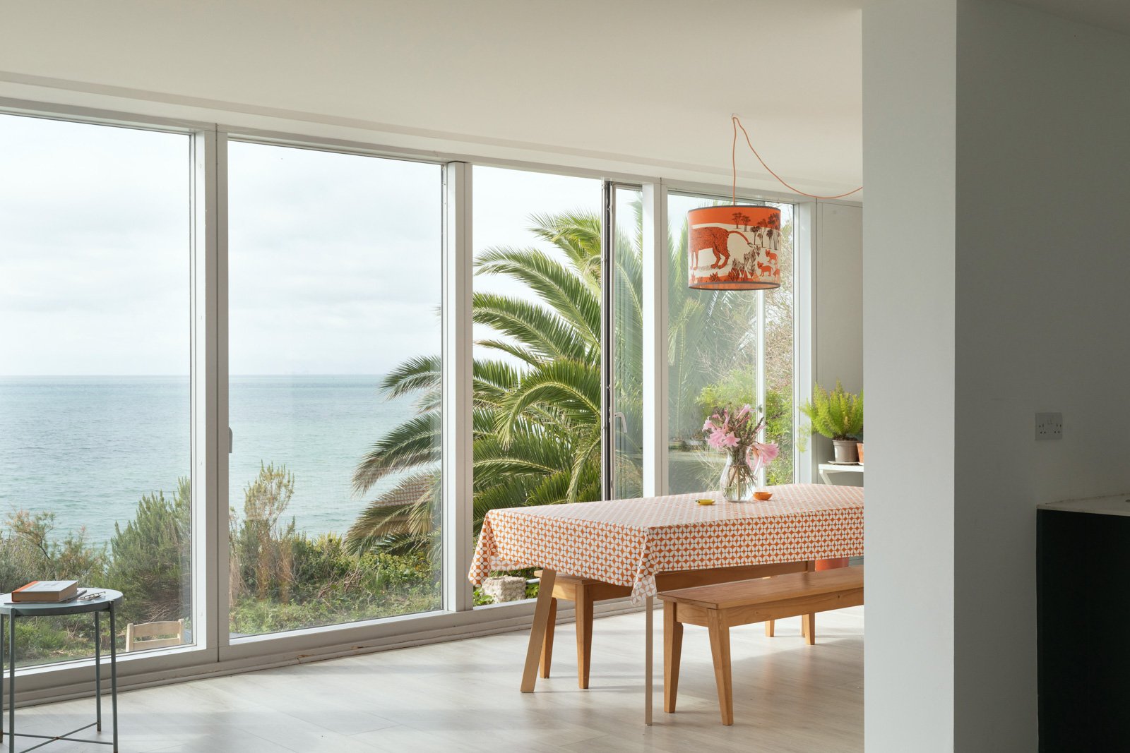 A bright living space with large window overlooking the sea and palm trees, with a table covered in an orange patterned tablecloth, a floral centerpiece, potted fern, and hanging lamps with safari animal design.