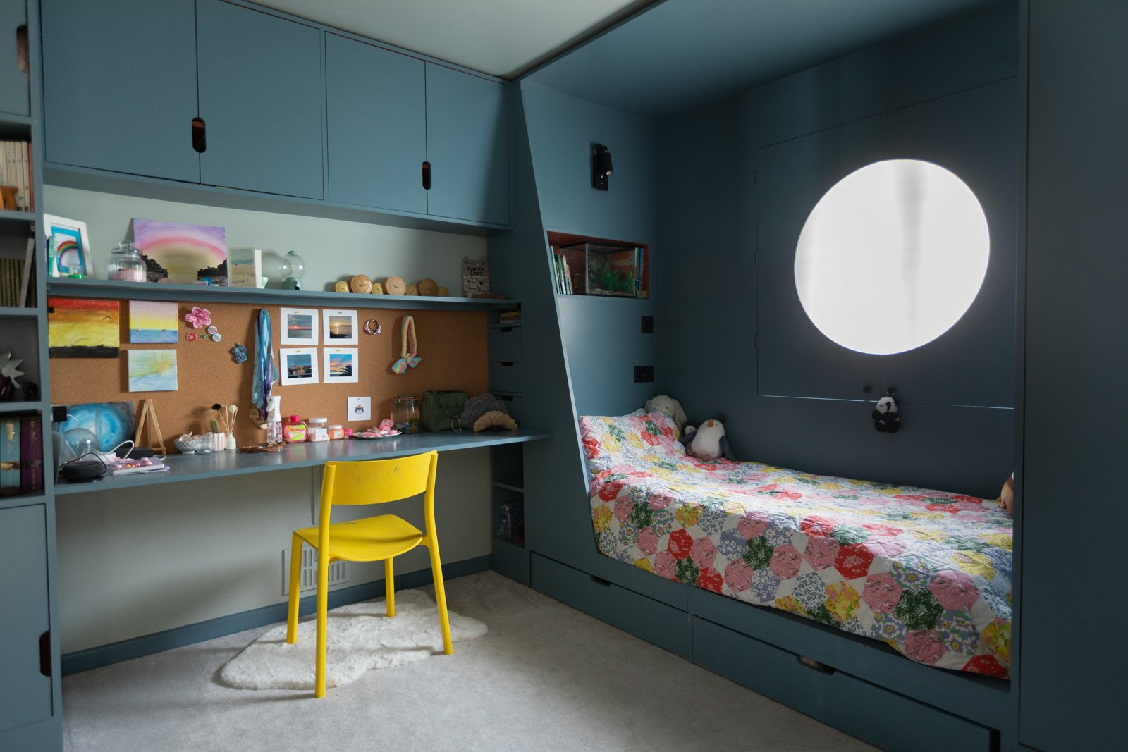 A cosy bedroom with a built-in bed featuring a colorful quilt, a window with a round opening, and a study area with a yellow chair, blue cabinets, and a corkboard decorated with artwork and photos.