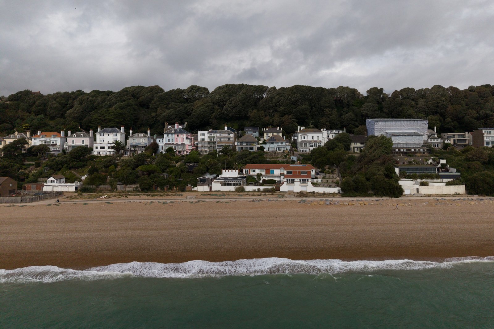 Beach with stones and gentle waves, overlooking a hillside filled with residential houses under a cloudy sky.