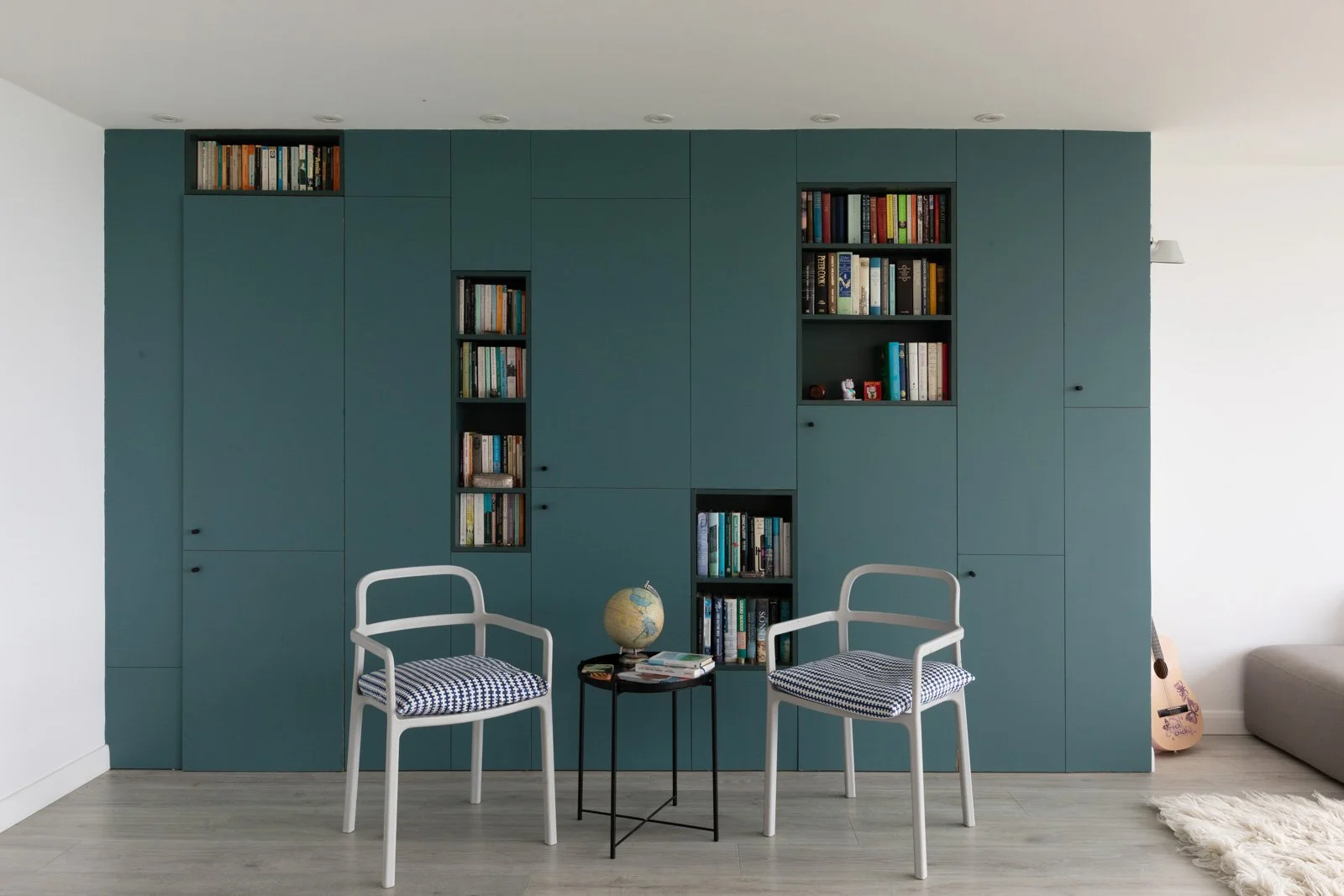 Living room with a large teal wall of cabinets and open shelves filled with books, two white chairs with striped cushions, a small black side table with a globe and books, a guitar leaning against the wall, and a beige sofa in the corner.