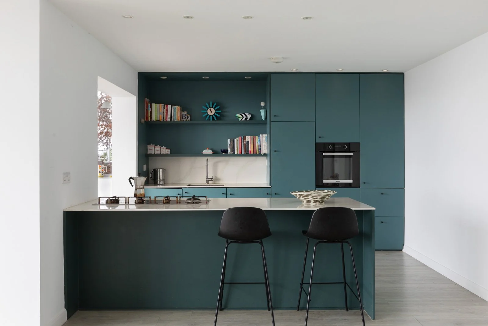 Modern kitchen with teal cabinets, white countertops, and a breakfast bar with two black chairs. Open shelves with books and decorative items behind the sink. Built-in oven, and outdoor view through a side window.