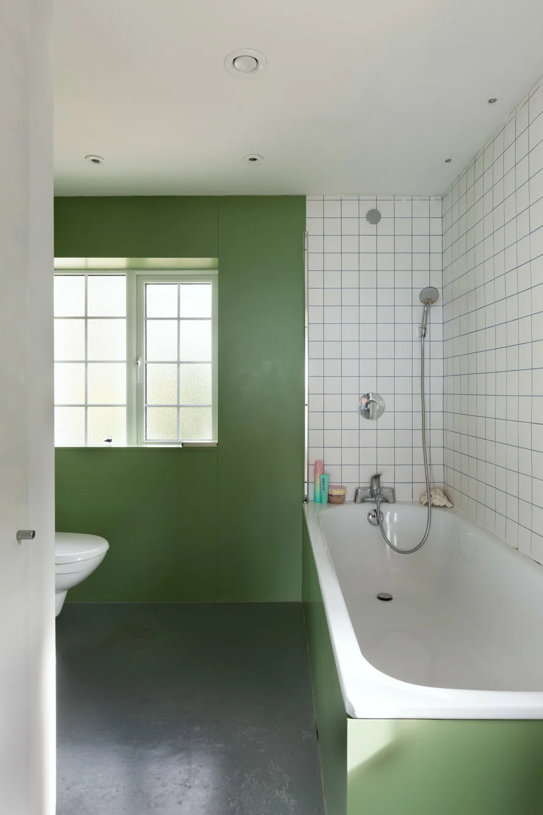 Bathroom with green walls, window, white toilet, white bathtub with showerhead, and toiletries on the tub edge, white tiled wall, and grey floor.