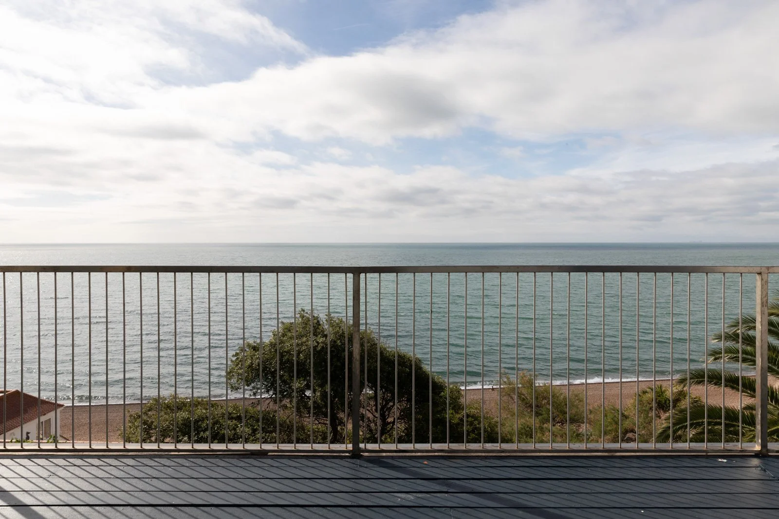 View of the sea through a balcony railing, with a partly cloudy sky and some trees and rooftops visible below.