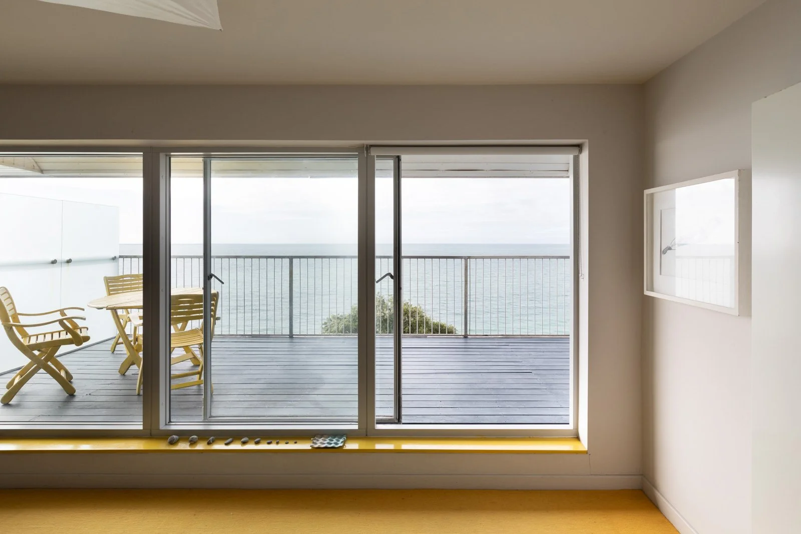 Interior view of a room with a large sliding glass door opening to a balcony overlooking the sea, with yellow outdoor dining furniture on the balcony.