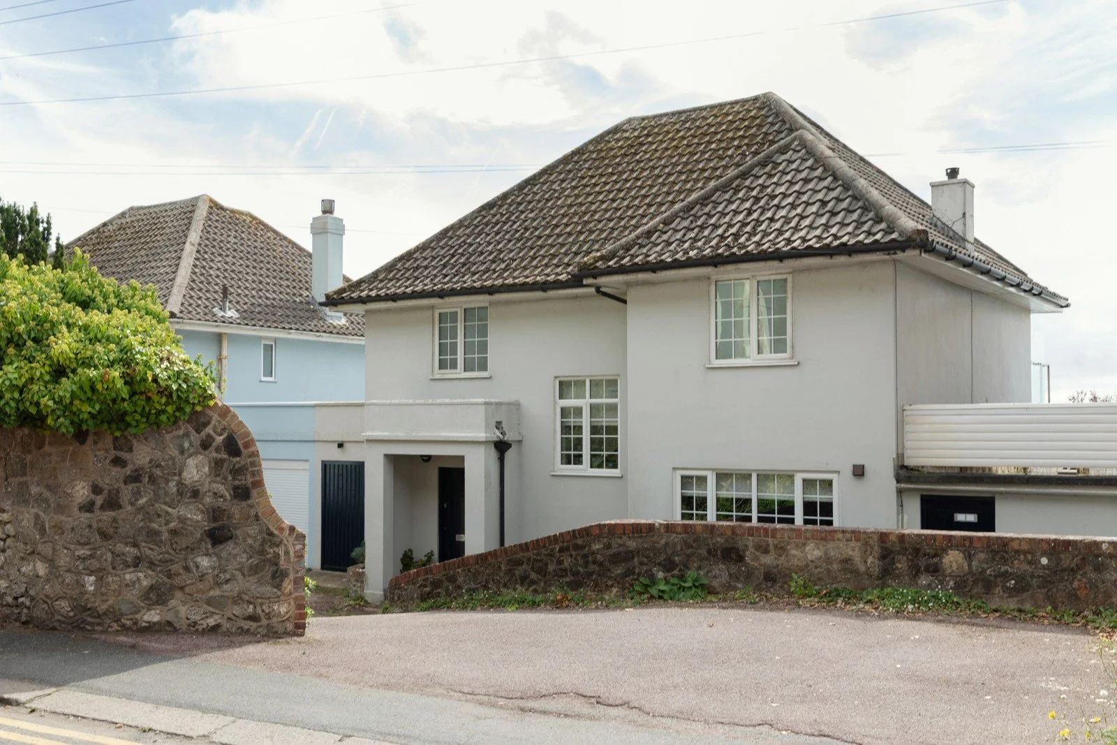 A white two-story house with a tiled roof, surrounded by a stone wall and greenery, on a suburban street.