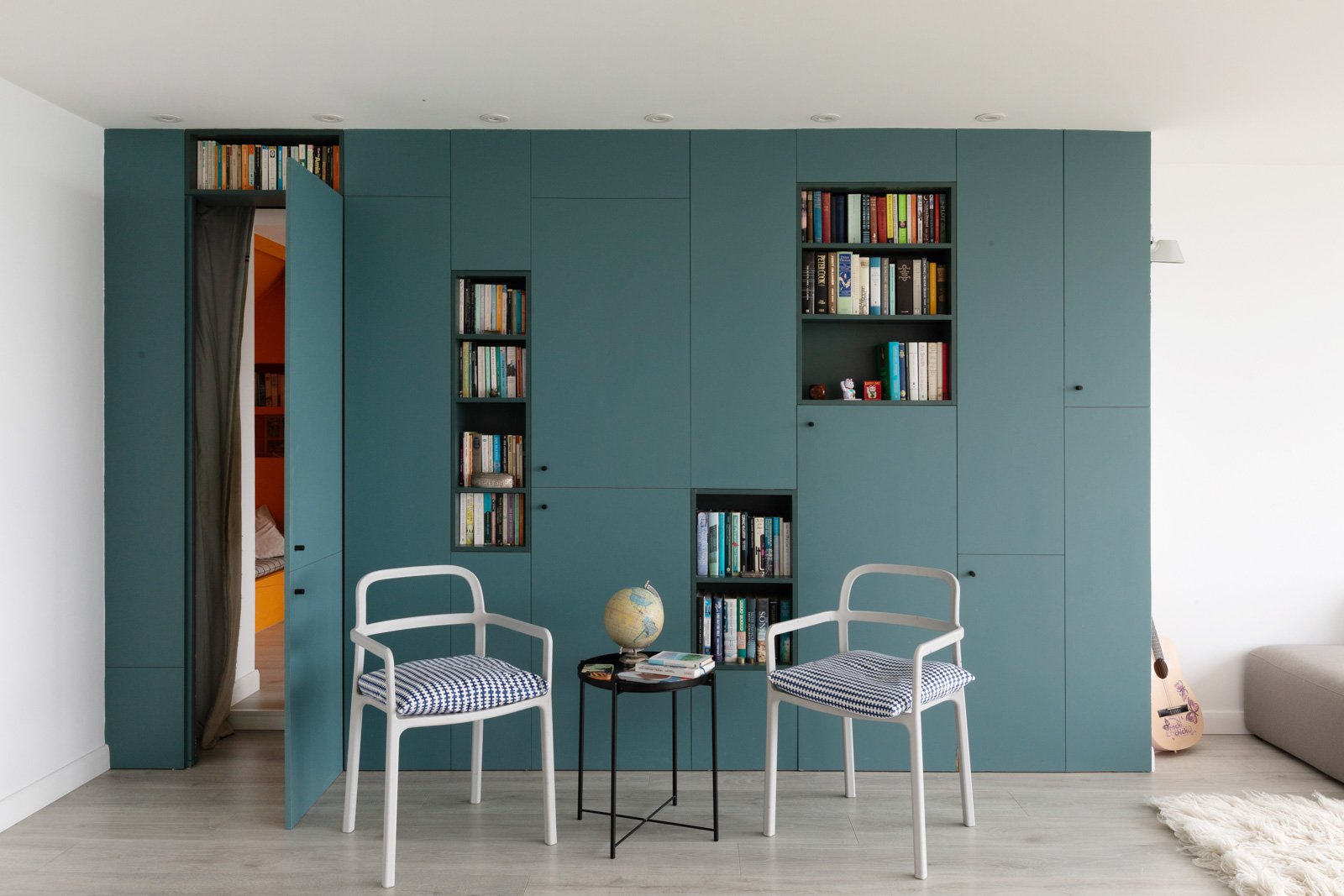 Living room with a large teal bookshelf wall divided into open cabinets filled with books, two white chairs with blue and white checkered cushions, a small black side table with a globe and books, a guitar leaning against the wall on the right, and a white furry rug on the light wood floor.