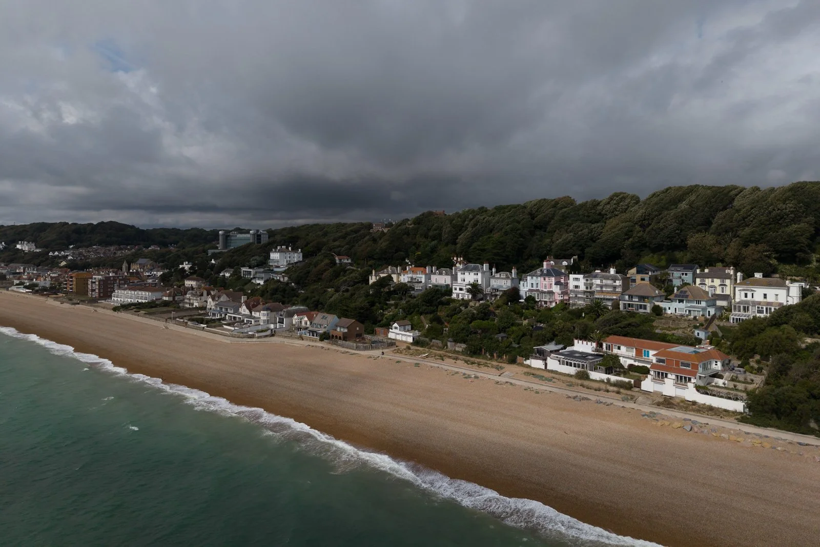 A coastal scene with a stoney beach in the foreground, ocean waves, and a hillside town with colorful houses and greenery under a cloudy sky.