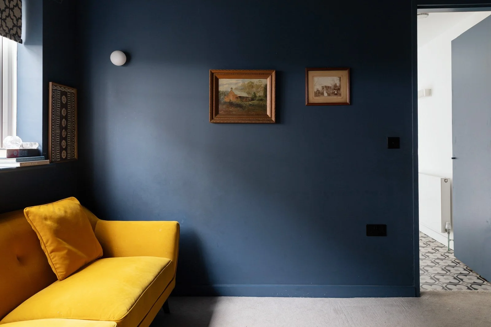 Living room with a yellow sofa against a dark blue wall decorated with two framed paintings, a window with a books and objects on the sill, and a doorway showing patterned floor tiles in an adjacent room.