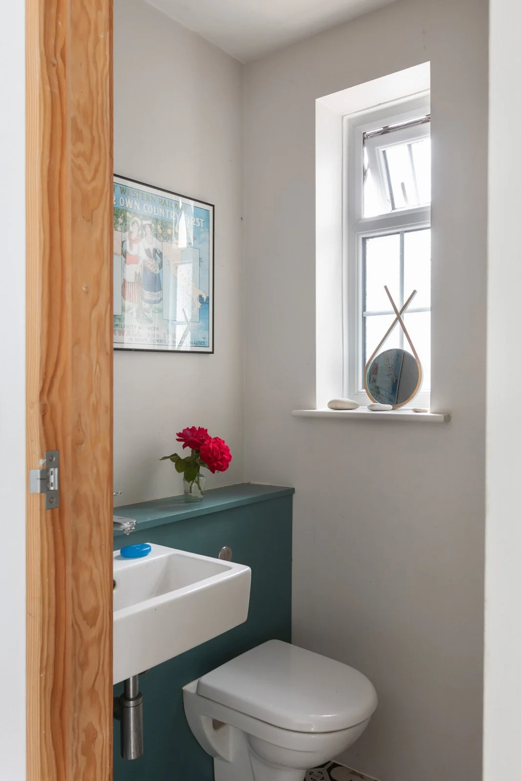 A small bathroom with a white wall, a window with a small mirror and decorative stones, a dark green cabinet with a vase of red flowers, and a framed picture on the wall.