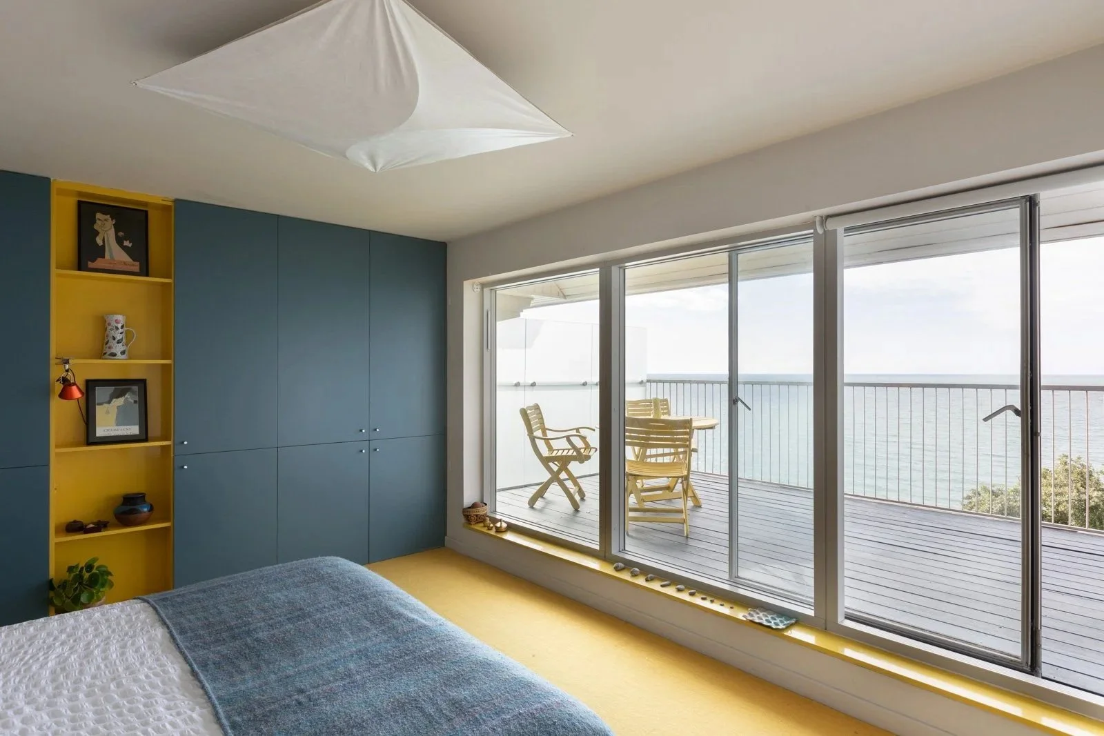 Interior view of a bedroom with a sliding glass door opening onto a balcony overlooking the sea, with a small wooden table and four chairs on the balcony.