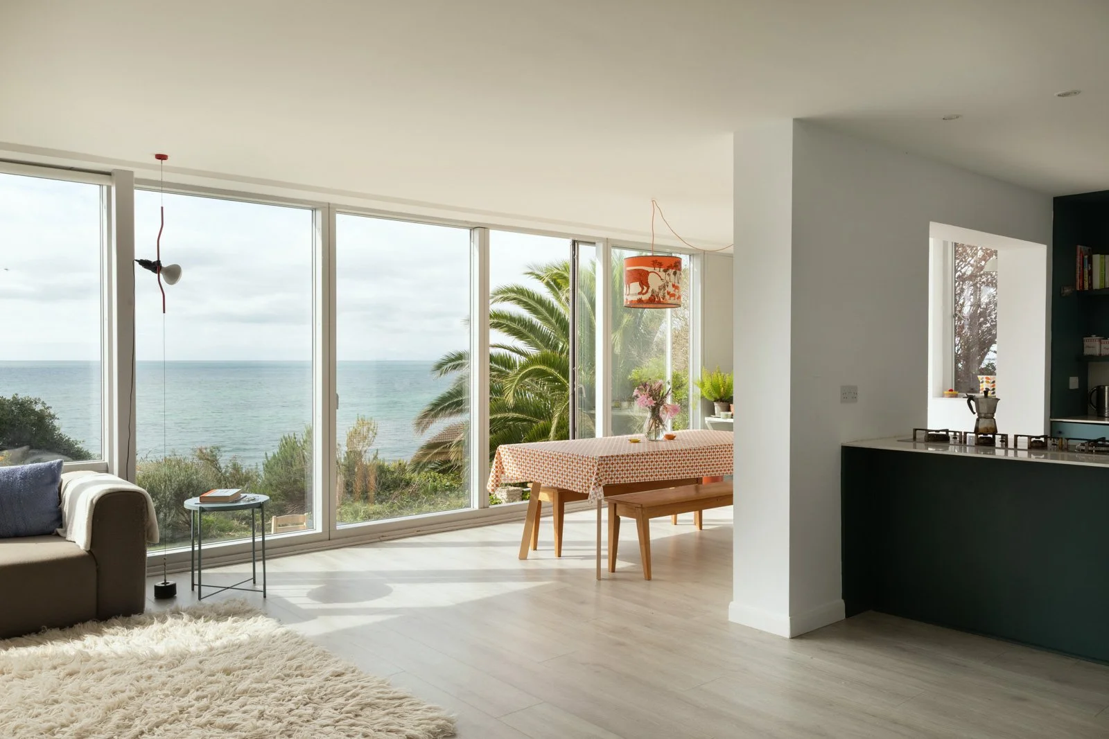 Living and dining area with large sea-view windows, a beige sofa with a pillow, a side table with a book, a shaggy rug, a dining table with a patterned tablecloth, plants, and a hanging lamp with a red shade, overlooking palm trees and the sea.
