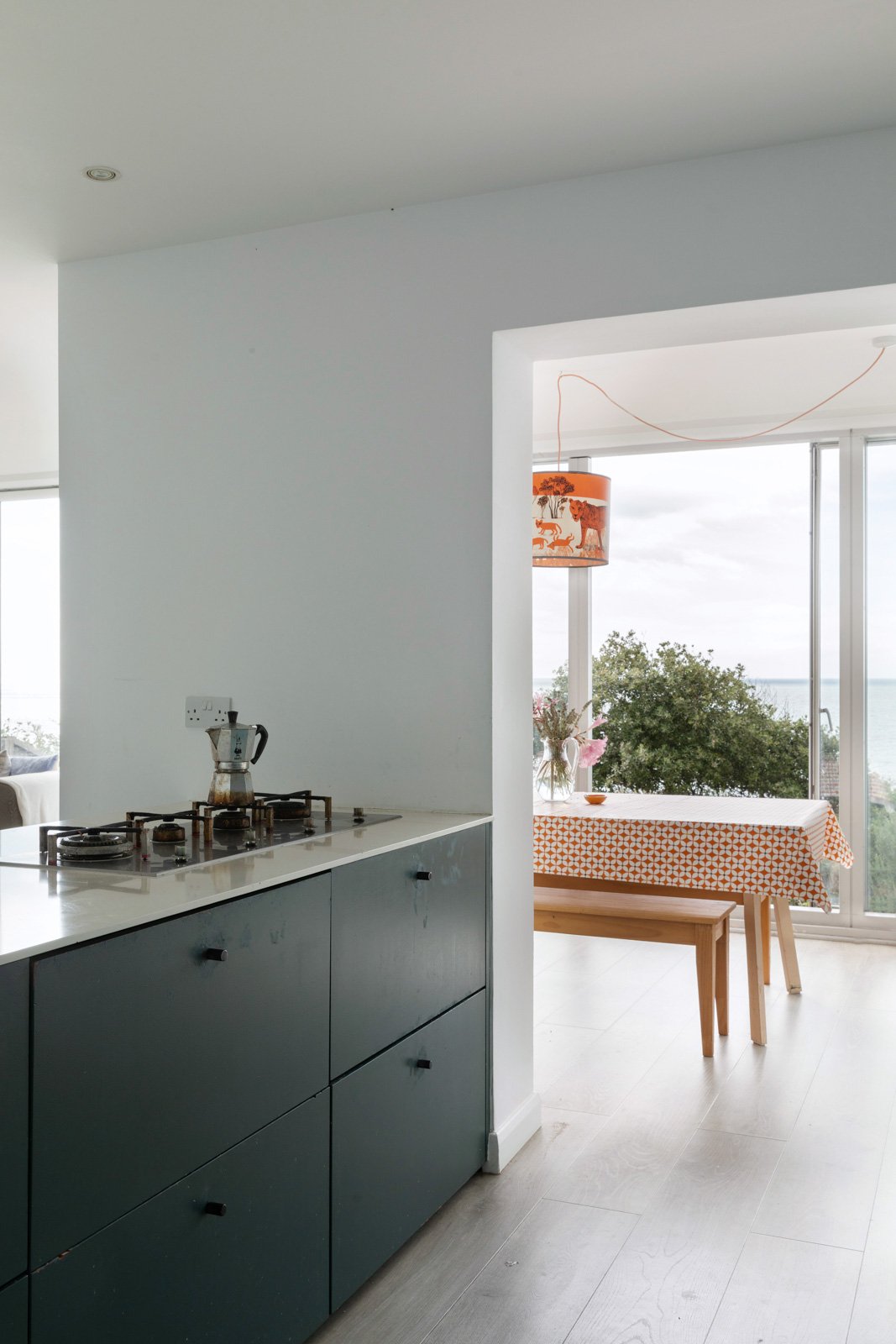 Modern kitchen with dark green cabinets, white countertop with stovetop and coffee pot, adjacent dining area with table, floral arrangement, patterned lamp, and large windows overlooking trees and water.
