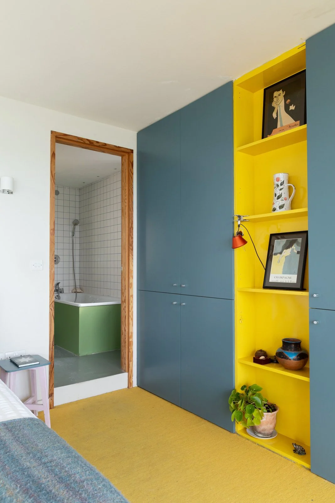 Interior of a room with a blue wardrobe, a yellow open shelving unit with decorations and framed photos, a small purple side table, and a doorway leading to a tiled bathroom with a green bathtub.