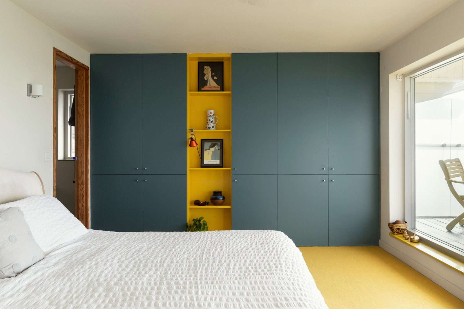 Bedroom with a blue wall unit, yellow shelves, bed with white quilt, window with a view, and decorated with artwork and pottery.